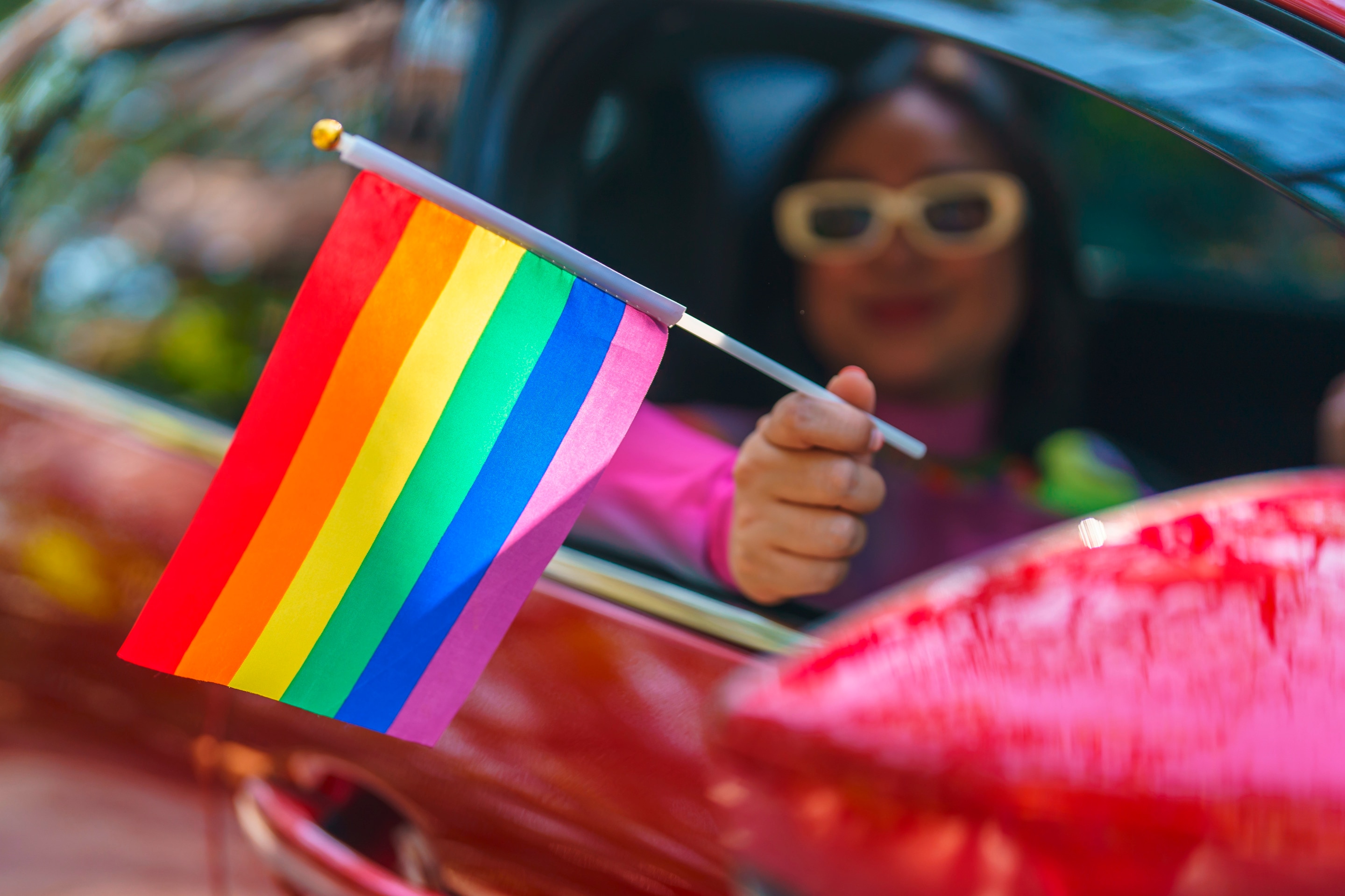 Asian queer woman in a car waving a Pride flag.