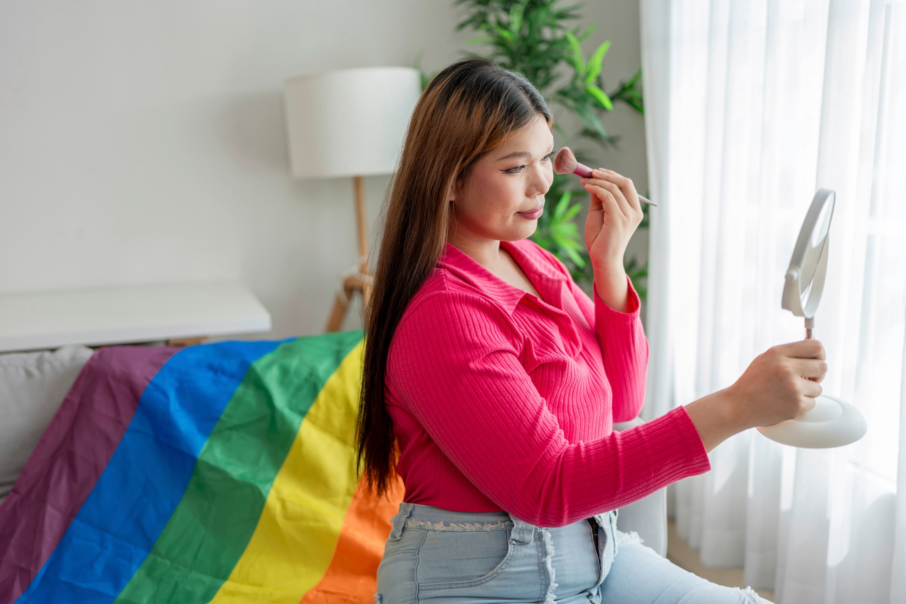 Asian transwoman applying makeup with a Pride flag in the background.