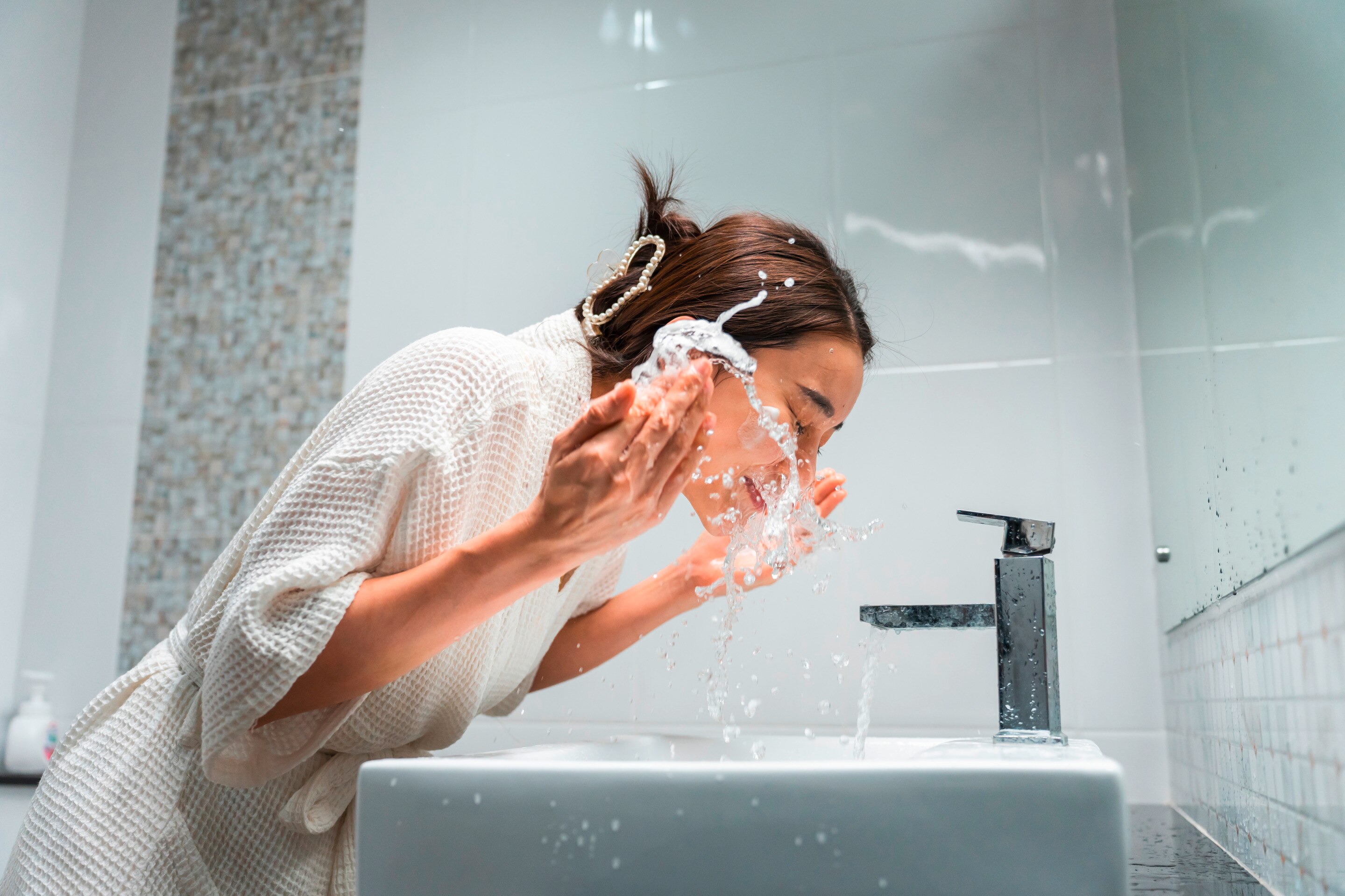 Woman in a robe washing her face with water.