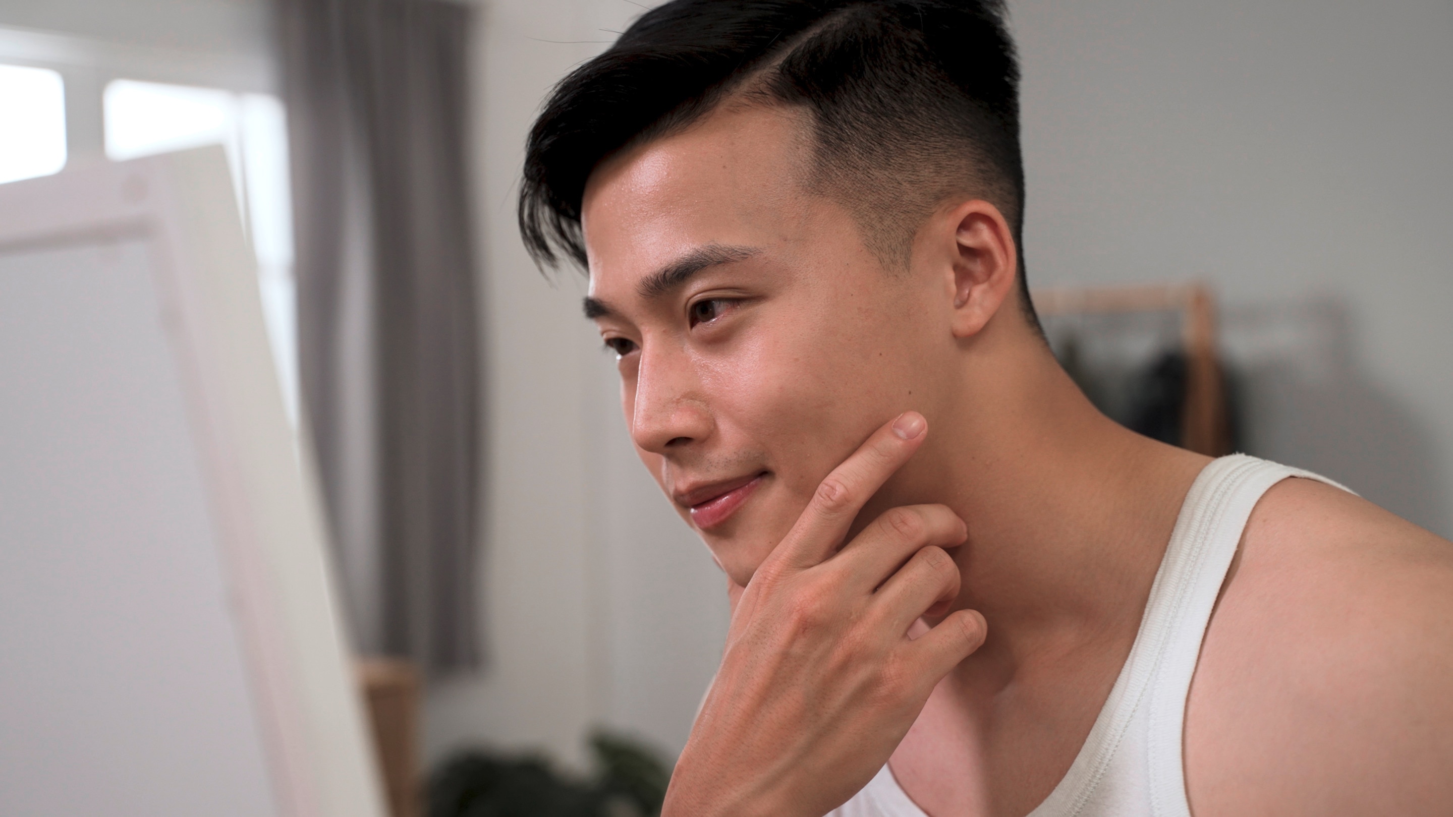 Man with short hair and white tank top looking at his face in the mirror.