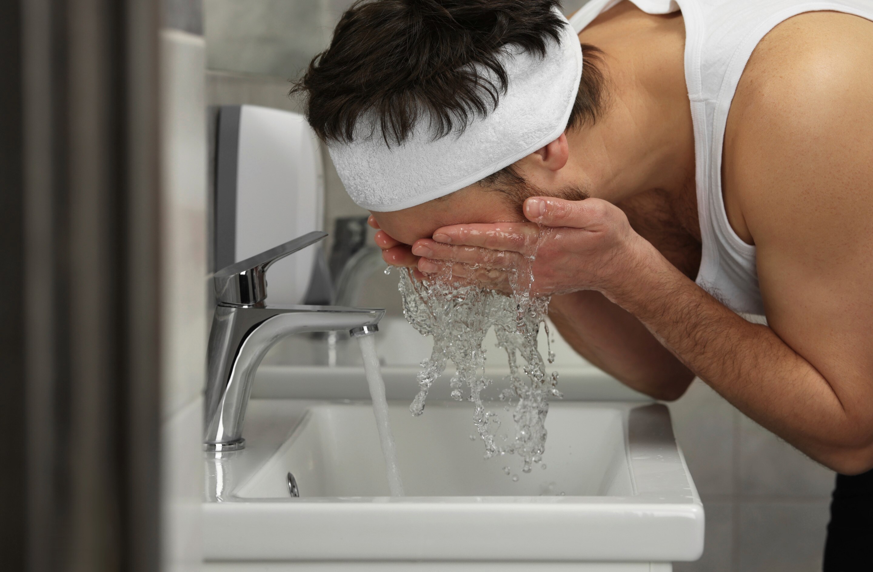 Man with white headband washing his face in the bathroom.