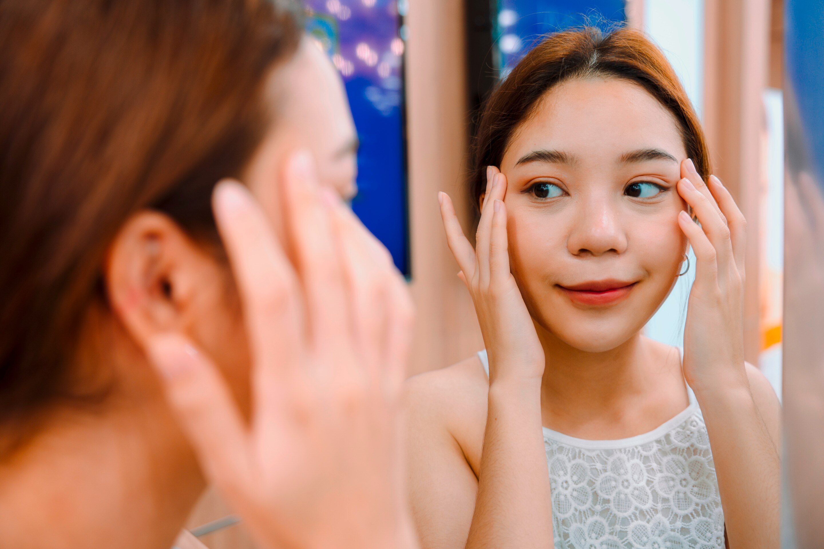 Woman with ponytail checking her under-eye area in front of a mirror.