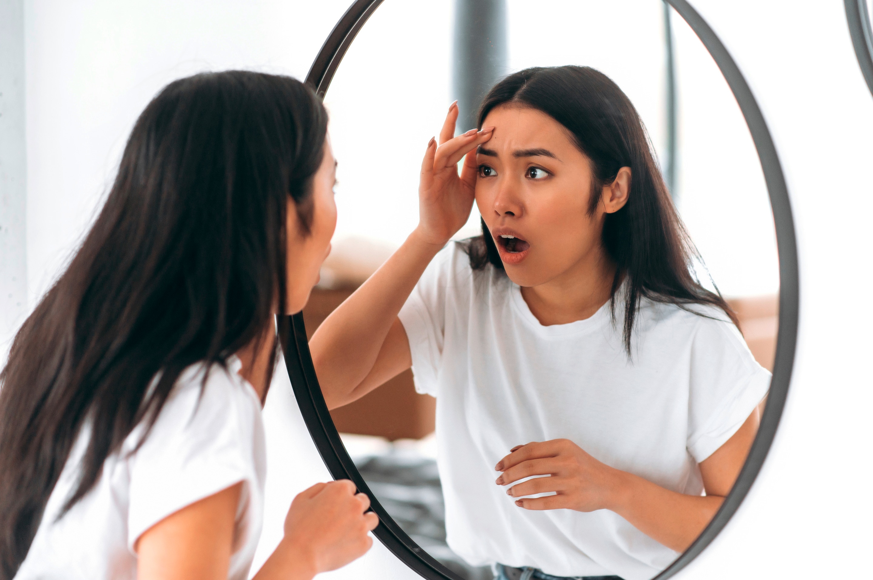 An Asian woman with a shocked expression looking in a mirror.