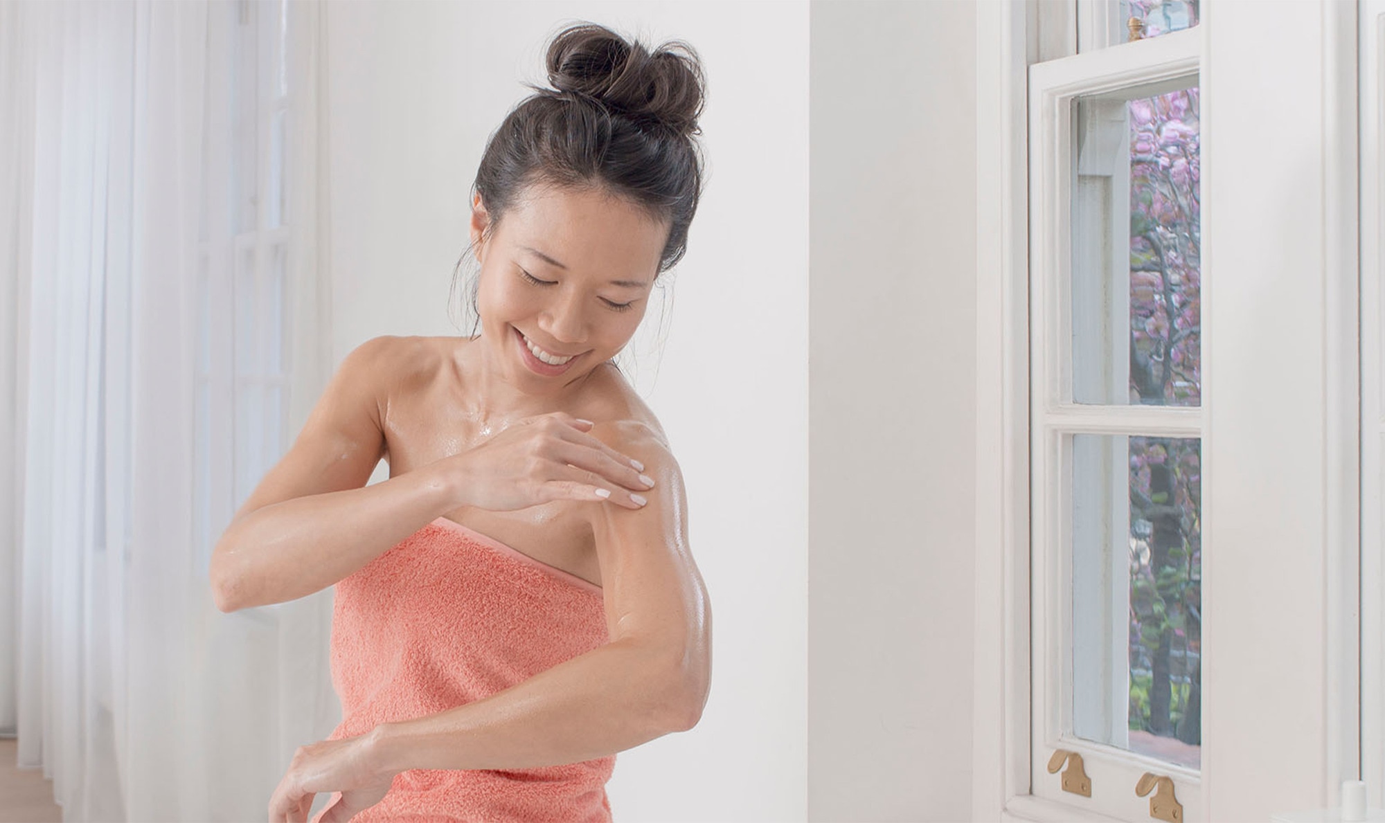 Smiling woman in pink towel applying moisturiser to her arm