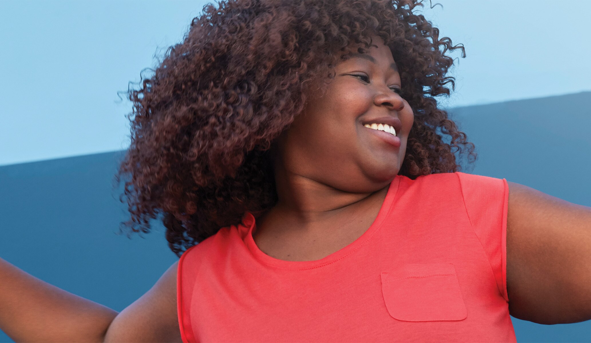 Image of woman smiling and dancing in salmon colored top against blue background