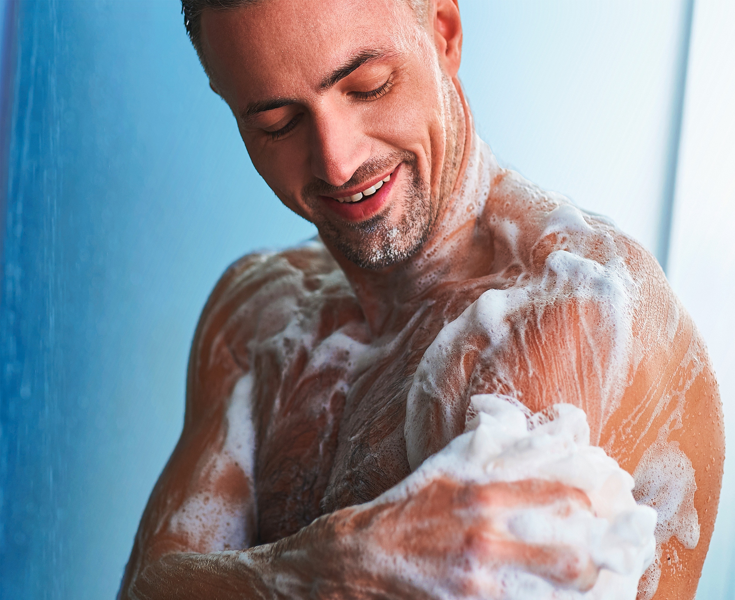 Man lathering body wash on his arm using a loofah in the shower