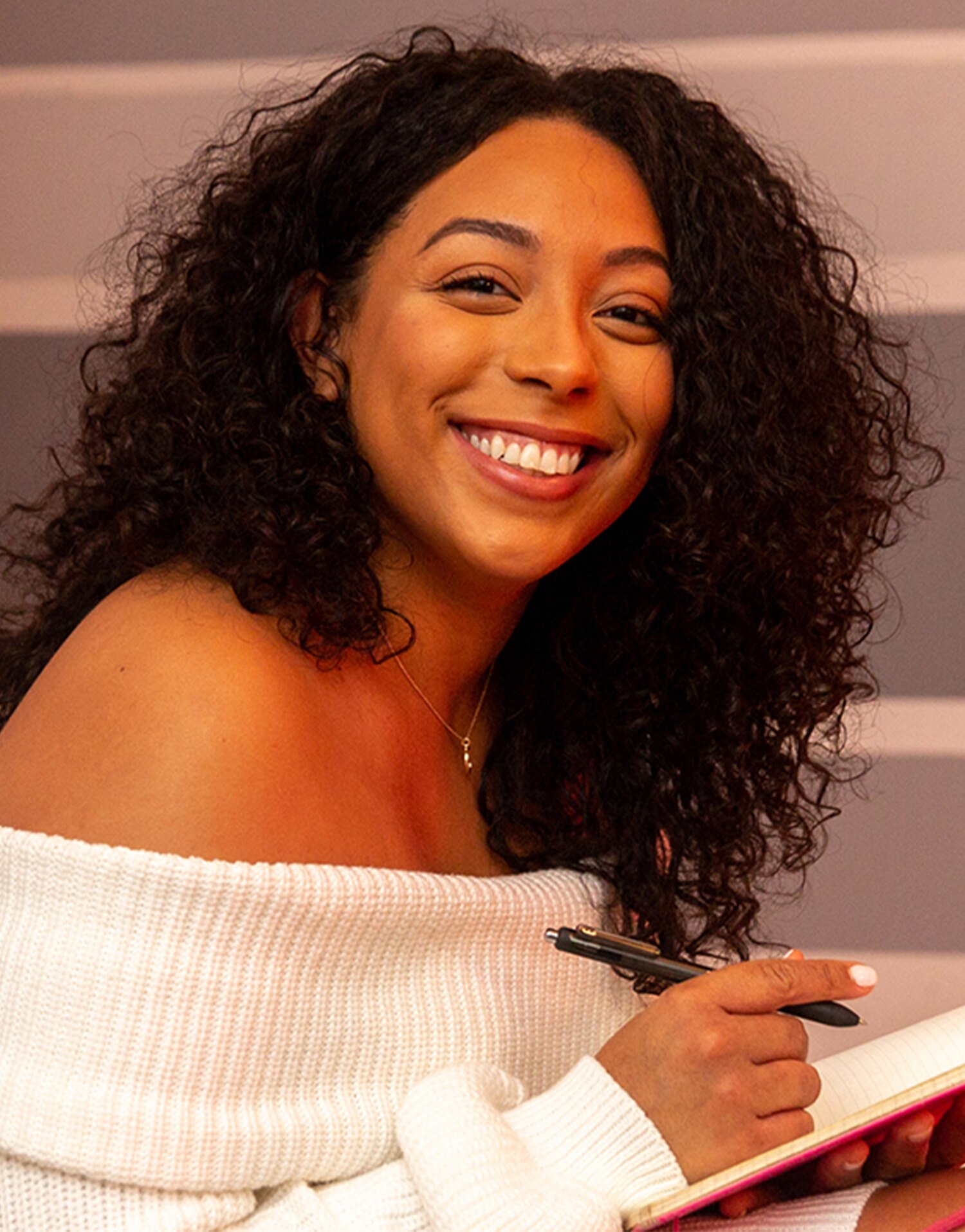 A woman smiles at the camera while writing into a notepad