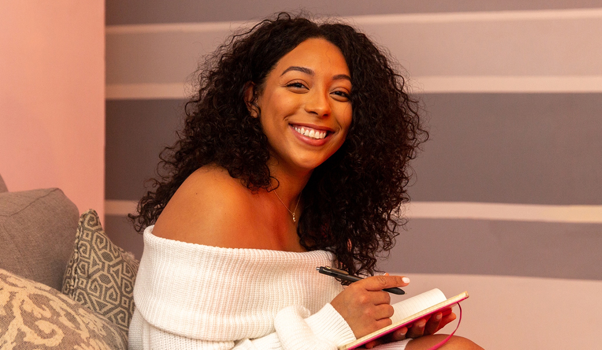 A woman smiles at the camera while writing into a notepad