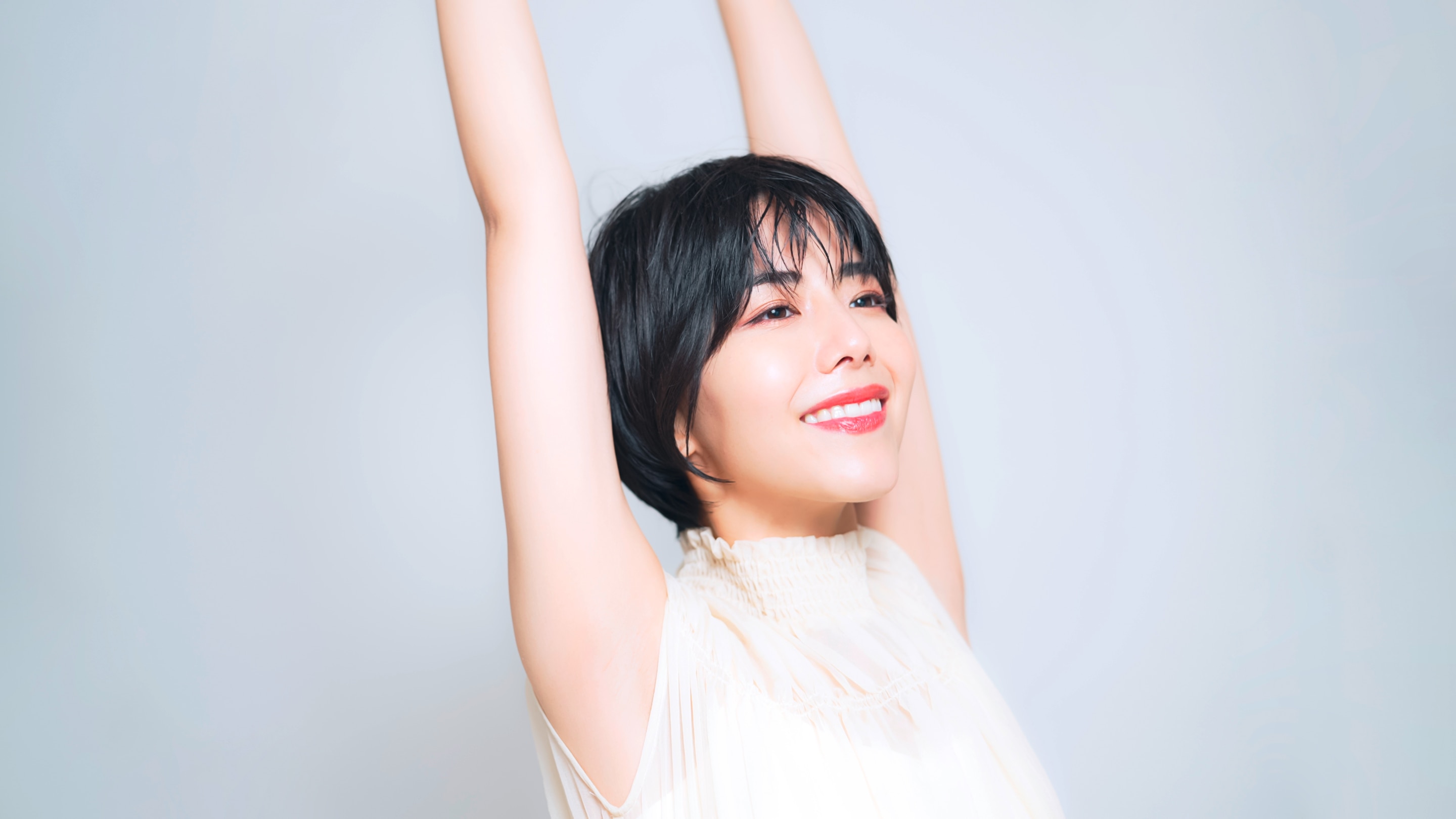 ผู้หญิงเอเชีย ผมสั้น ชูแขนสองข้าง ยิ้มมั่นใจ สวมเสื้อแขนกุดสีครีม