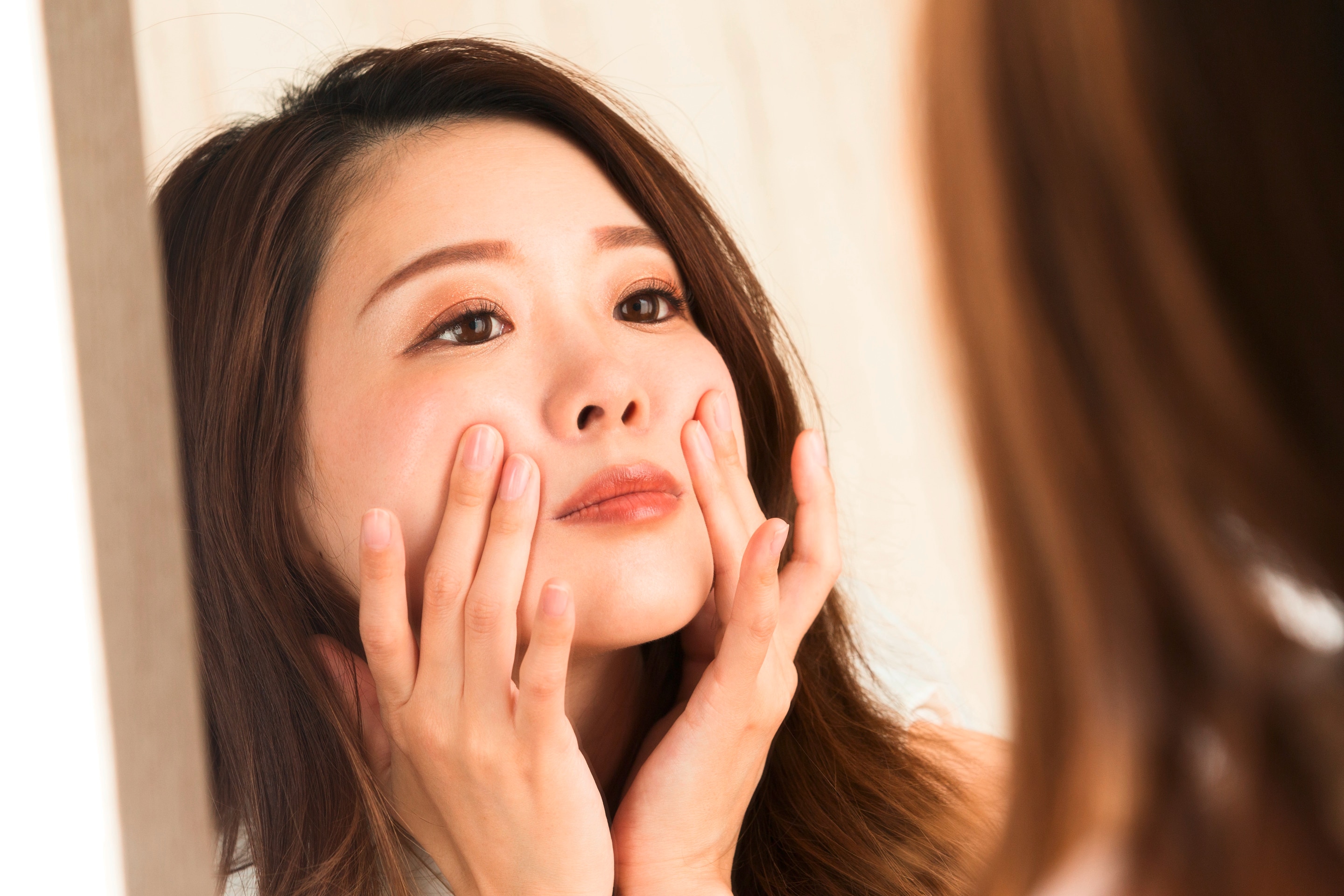 ผู้หญิงเอเชีย ผมแสกข้างยาวประบ่า ส่องกระจก สองมือแตะกดบริเวณร่องแก้ม