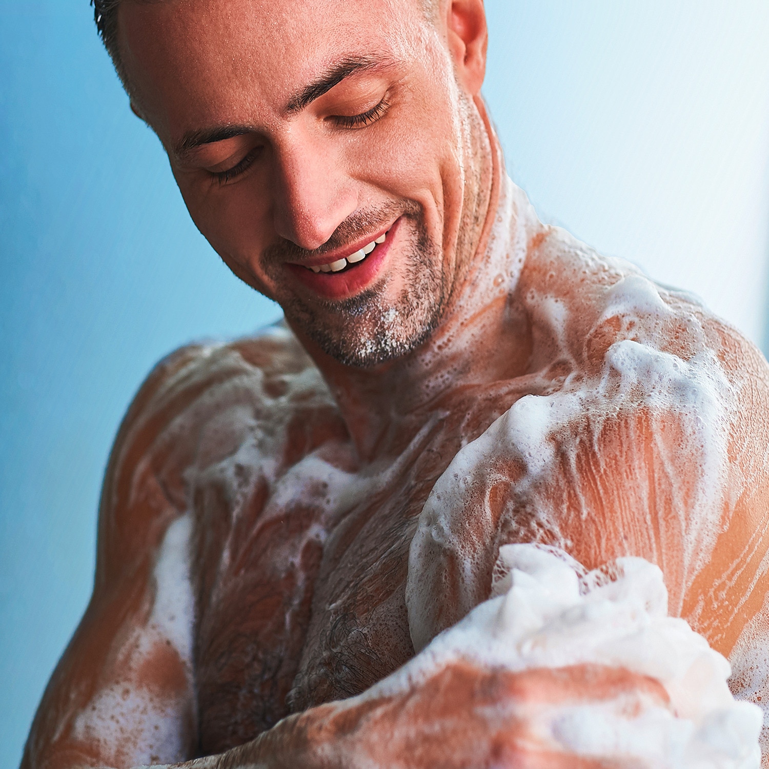 Man lathering body wash on his arm using a loofah in the shower