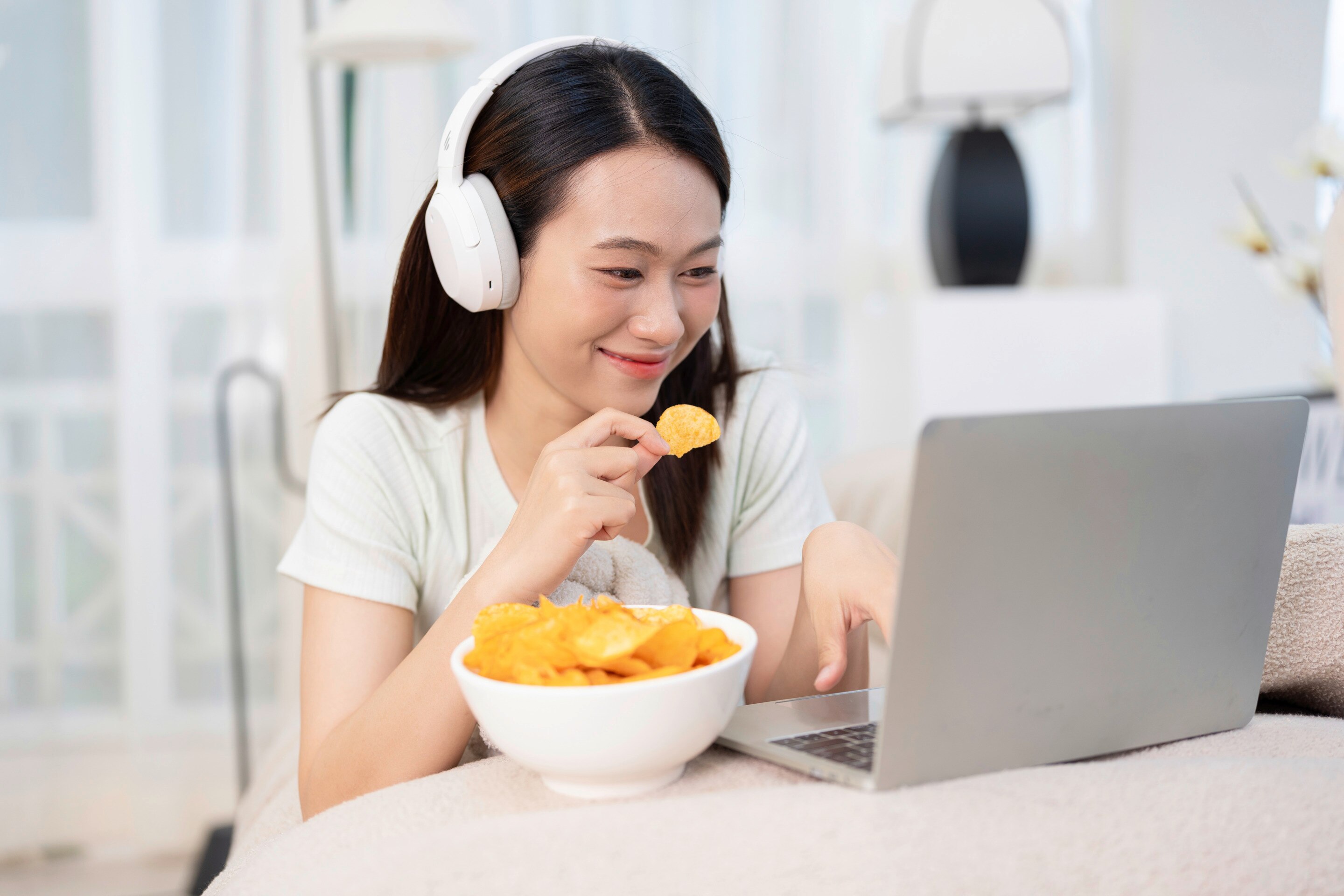 An Asian woman eating chips while working on her laptop.