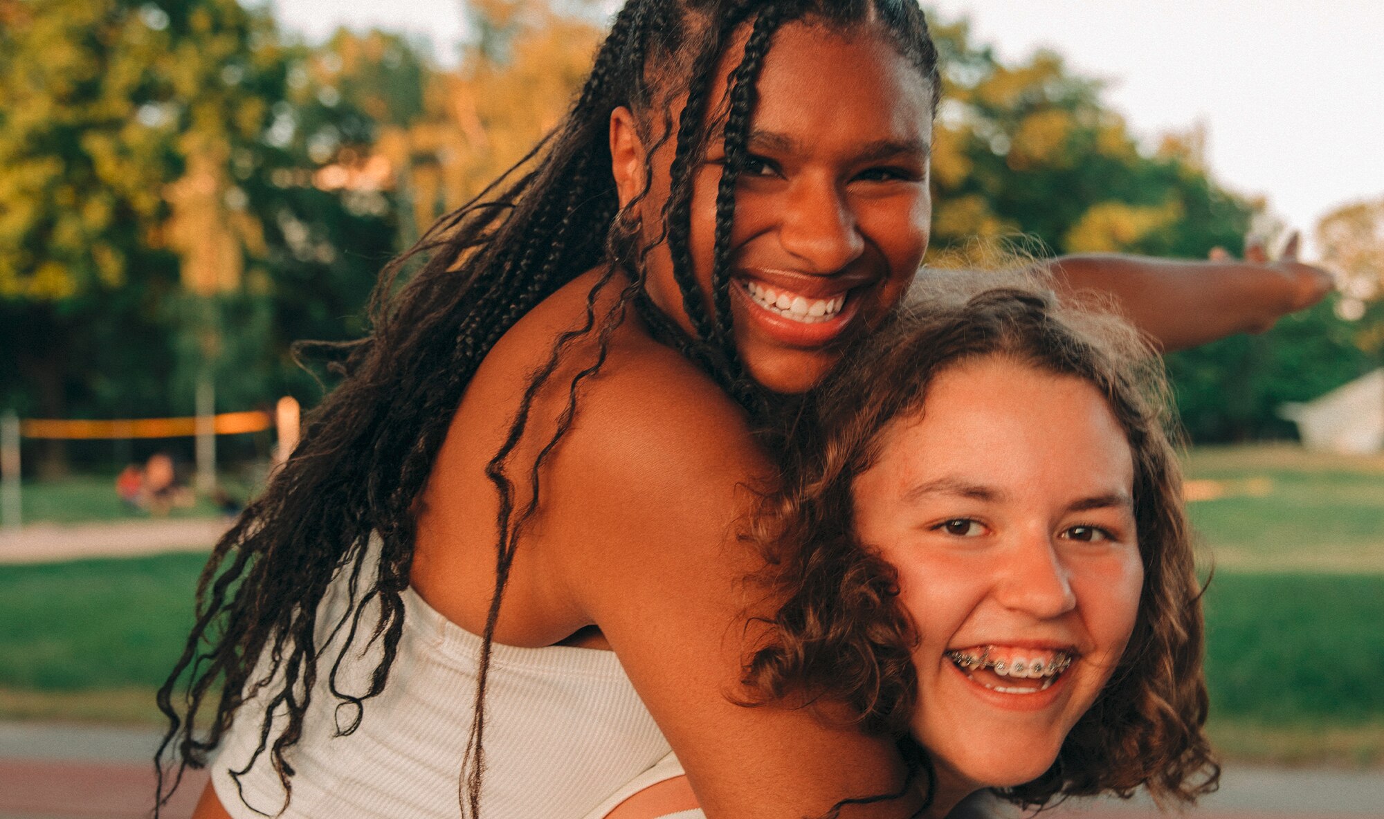 Two girls in a park, one is carrying the other on her back and they are smiling