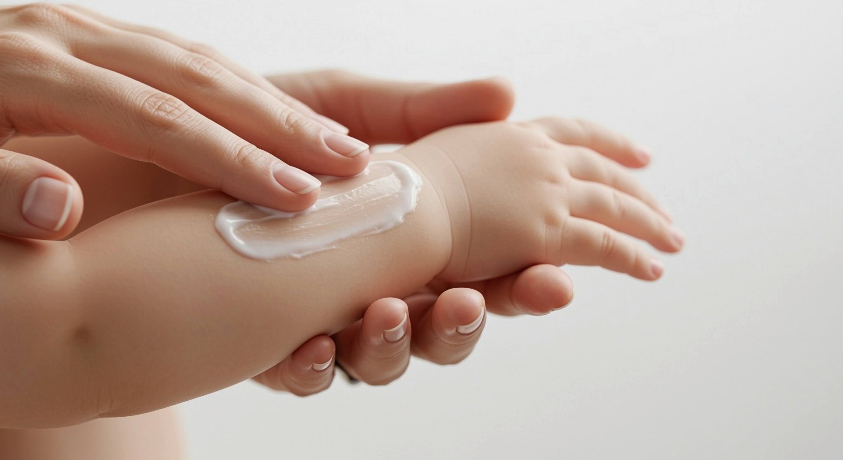 tangan orang dewasa mengoleskan krim hypoallergenic pada kulit lengan bayi untuk membantu menjaga kelembapan dan kenyamanan kulit bayi yang sensitif.