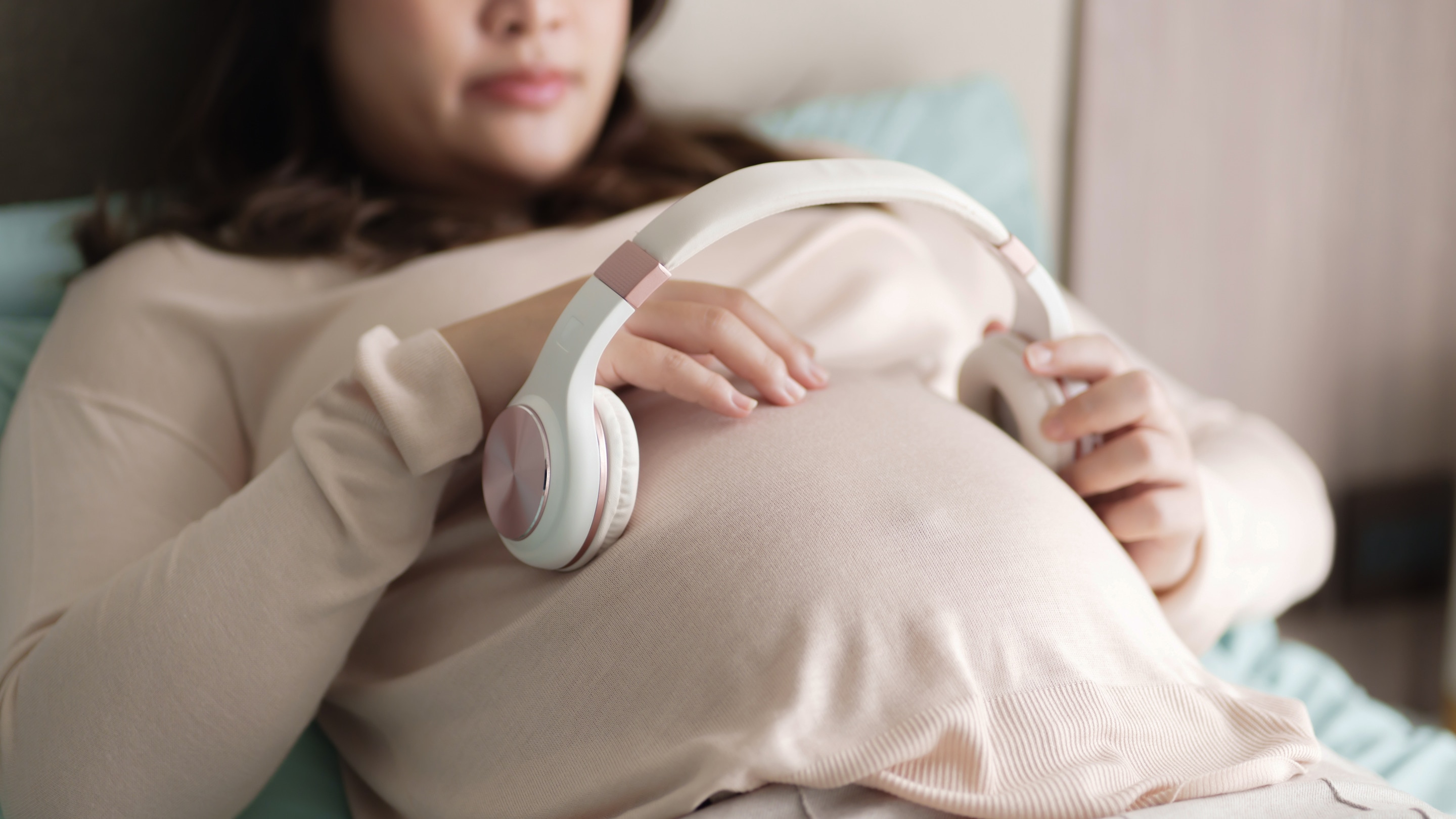 ada Ibu hamil yang sedang berbaring sambil mengenakan headphone di atas perut untuk melakukan stimulasi janin dalam kandungan melalui musik lembut.