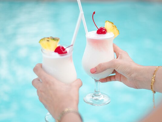 Female hands holding two Virgin Piña Colada Mocktails next to a swimming pool