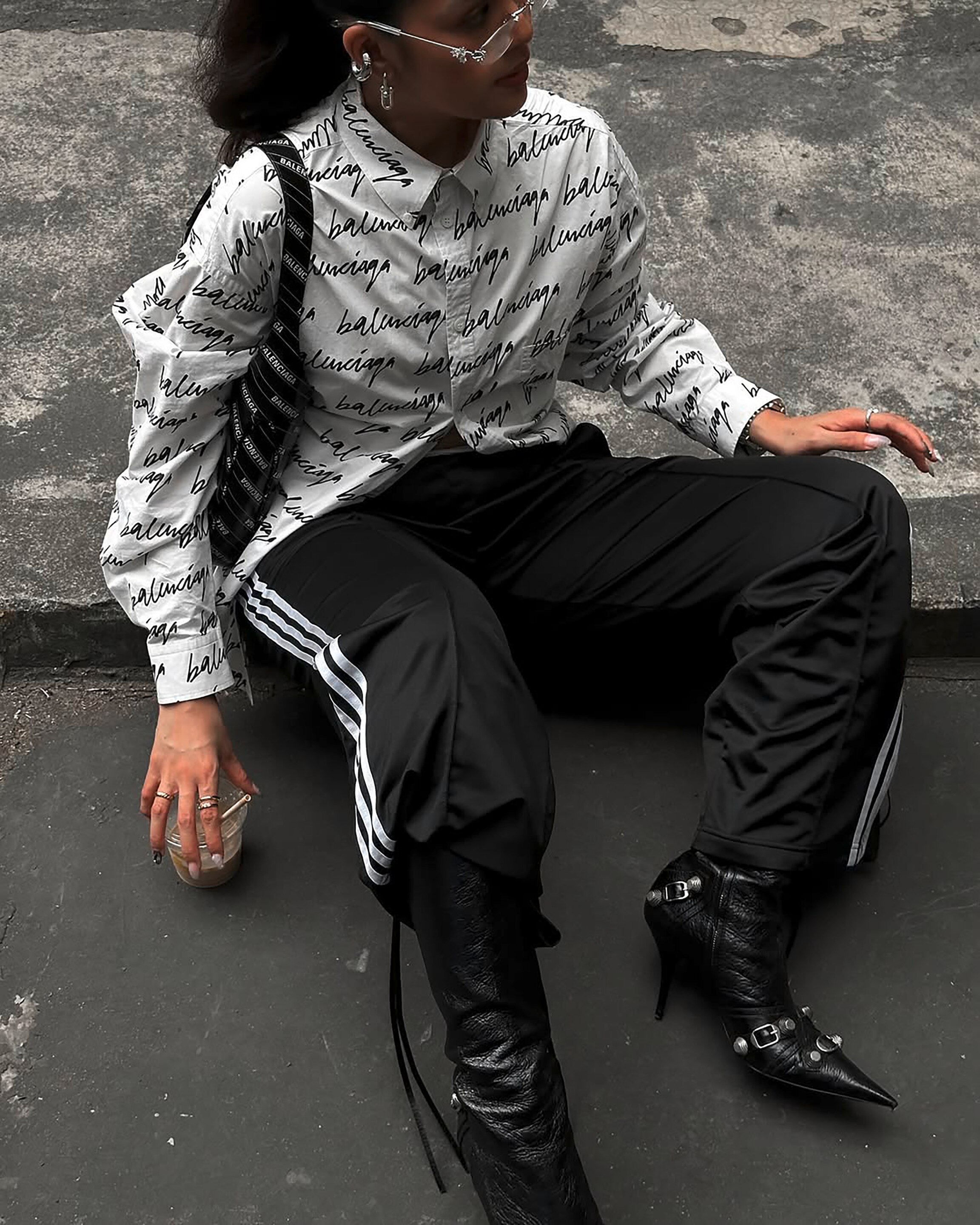 Fashionable woman wearing a button-down, tracksuit pants, and pointy-toed boots, sitting on the sidewalk.