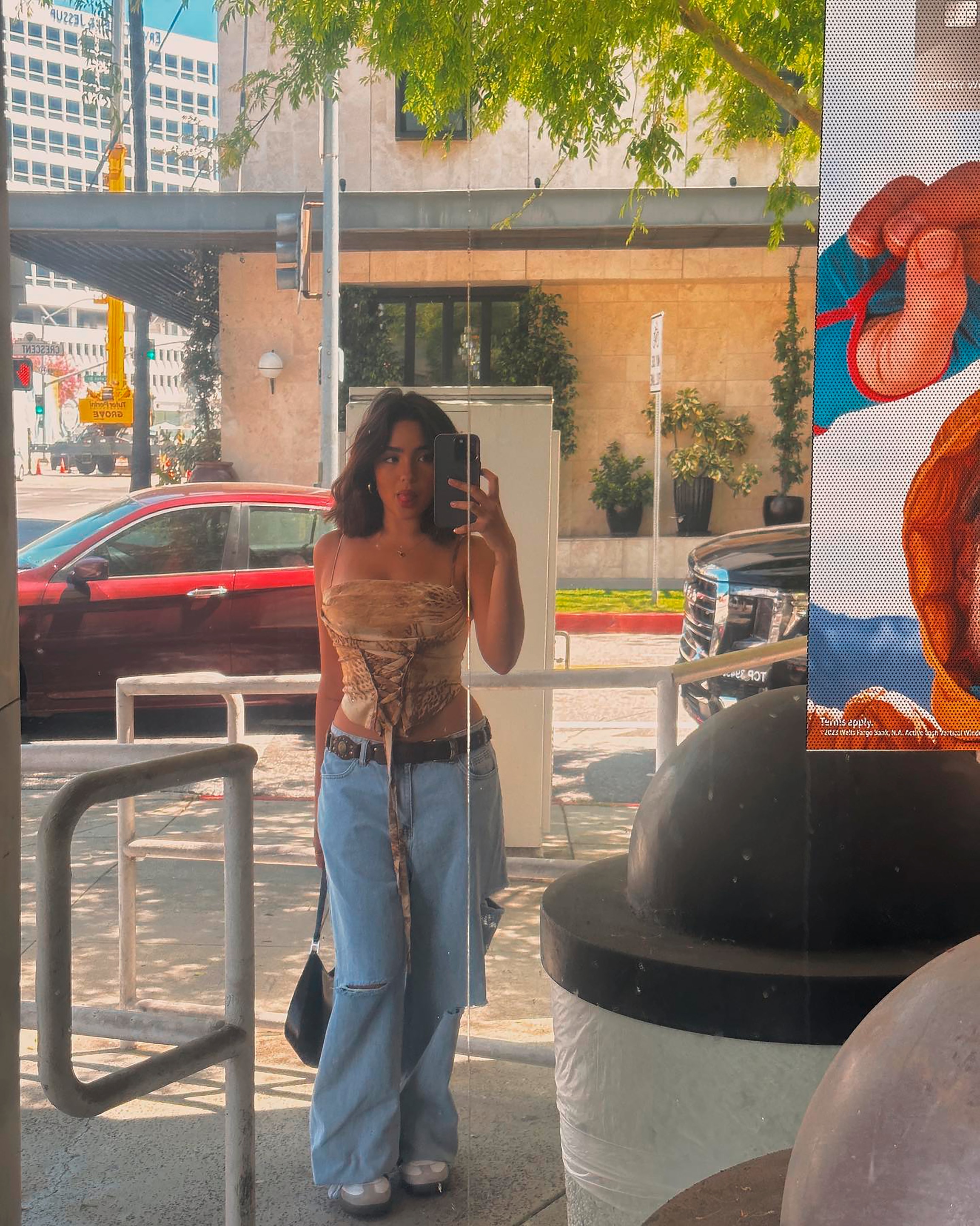 Woman in a corset top and baggy jeans, taking a full-body camera selfie outdoors.