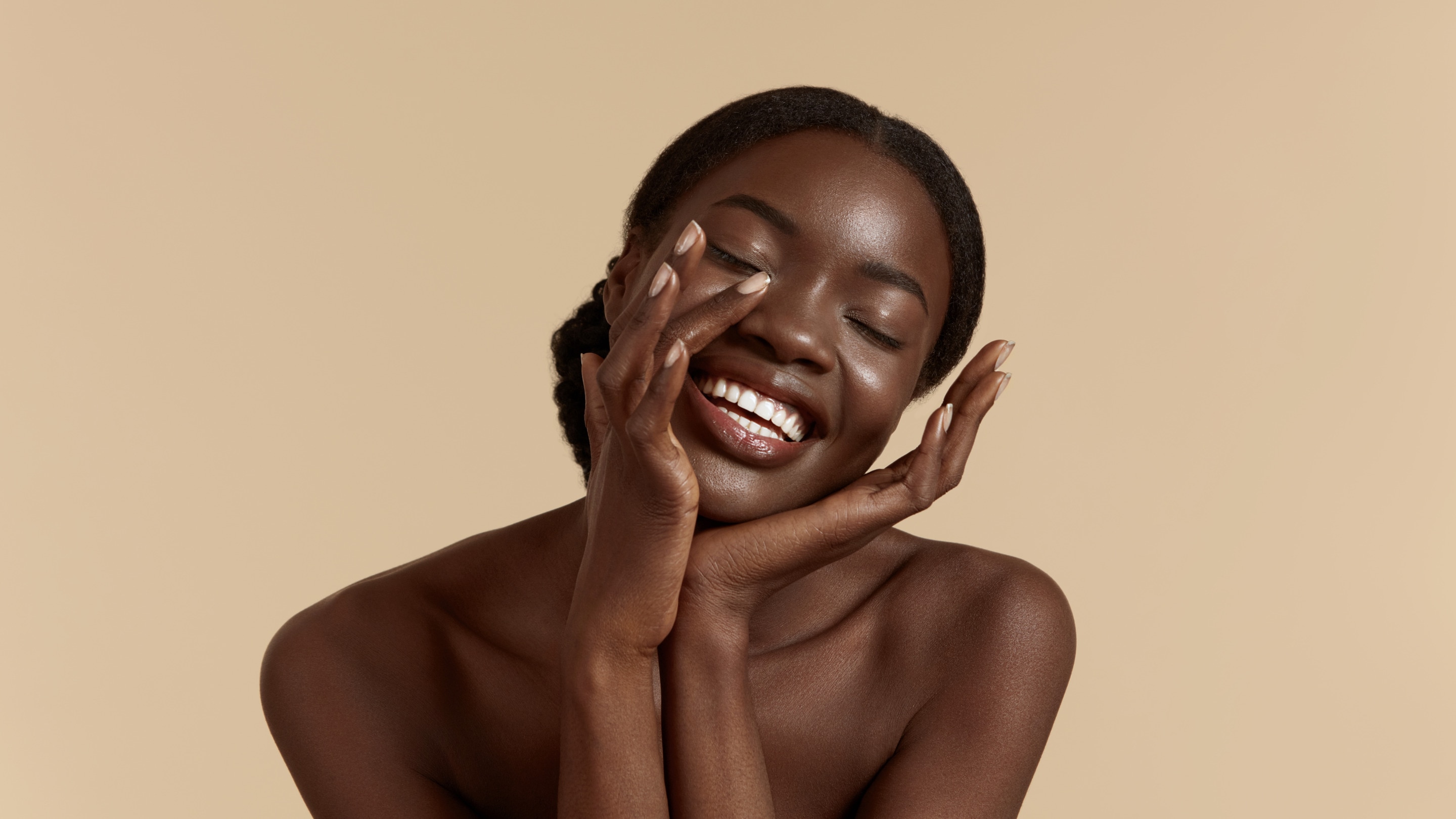Close-up portrait of a smiling woman with deep, radiant skin tone, eyes closed, gently touching her face with both hands against a neutral beige background.