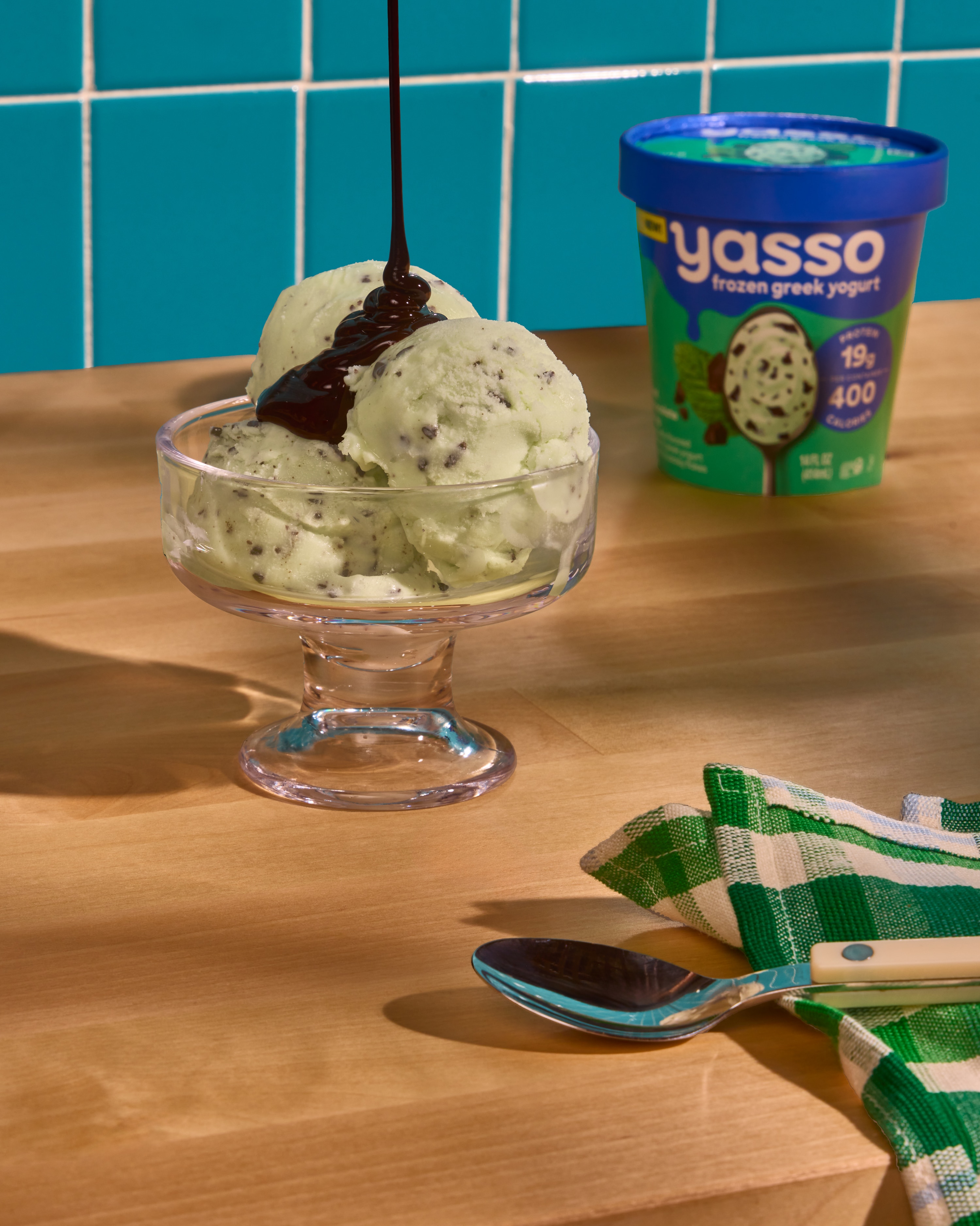 Scoops of Mint Chocolate Chip sit in sundae cup, Pint edges in background. Fudge drizzles from top of frame into cup.