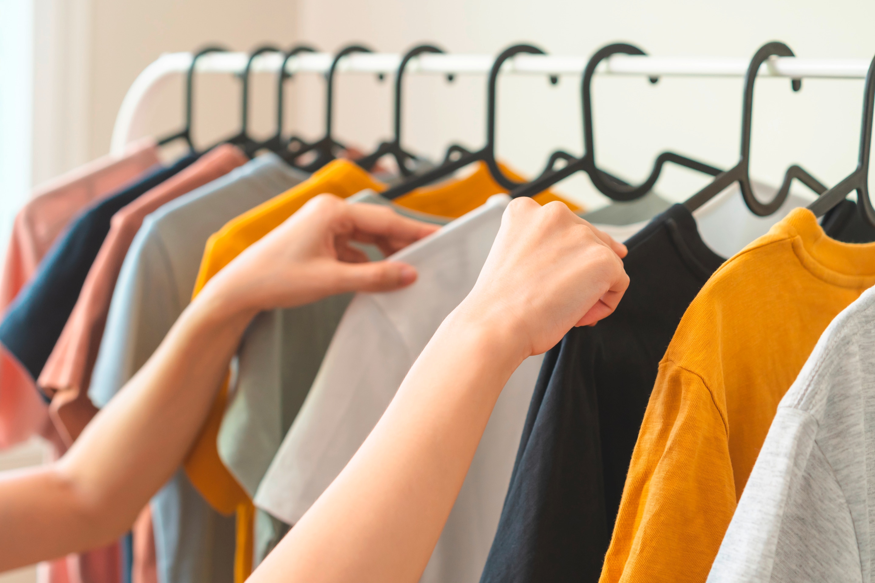 tangan seseorang sedang memilih kaos dari rak gantungan pakaian dengan berbagai warna.