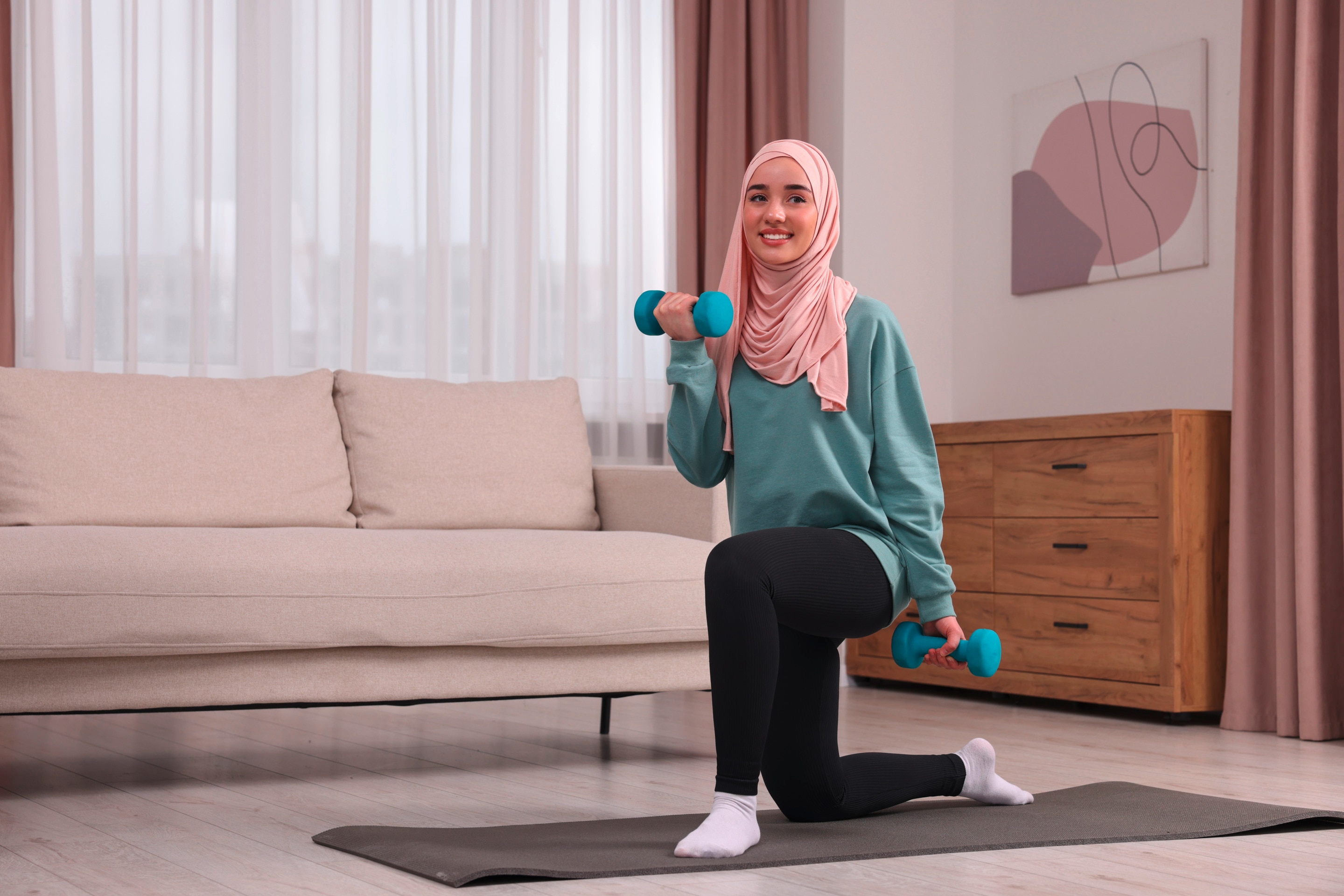 wanita berhijab sedang berolahraga di rumah sambil memegang dumbell.