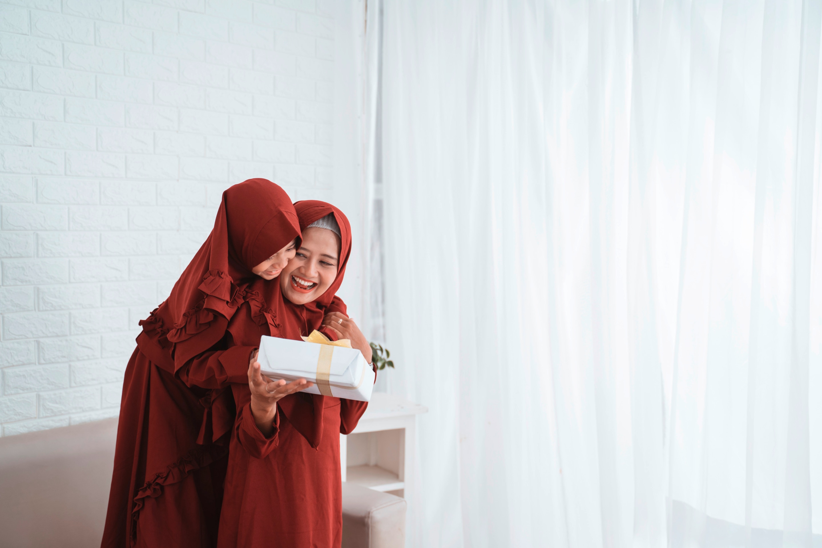 ada ibu dan anak perempuan berhijab mengenakan busana merah saling tersenyum sambil memegang hampers Lebaran.