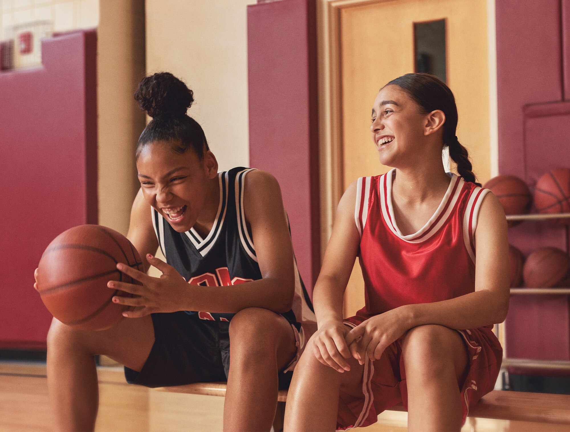 Two girls seated on a bench in a gymnasium laughing, one has a basketball in her hand