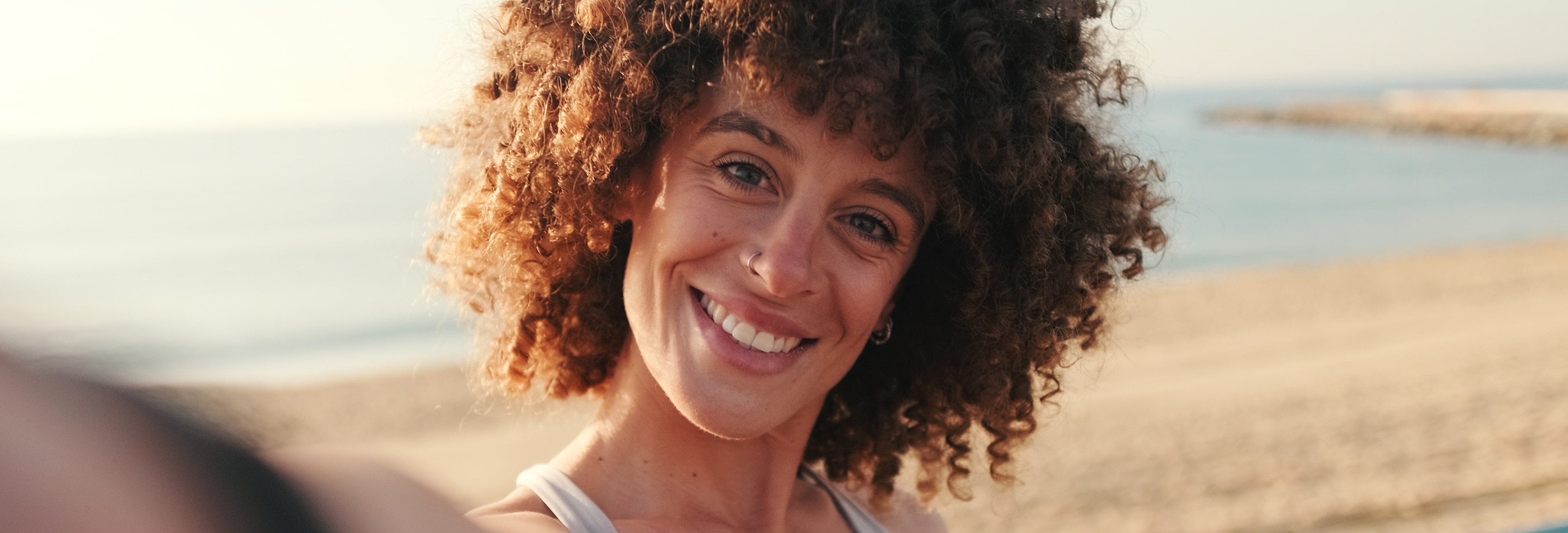 Woman with tight brown curly hair smiling with sunny beach and ocean in the background. 