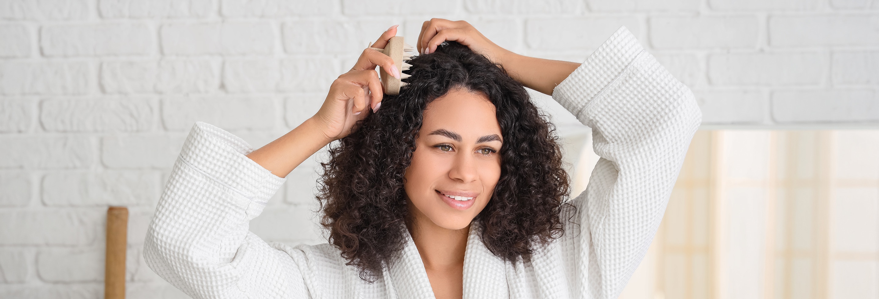 A woman in a white bathroom with dark curly hair gently combs her scalp