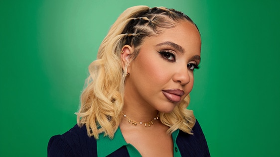 A woman with a silk press hairstyle, featuring sleek, straightened blonde hair with cornrow braids along the crown, posing against a green background wearing a dark blazer.