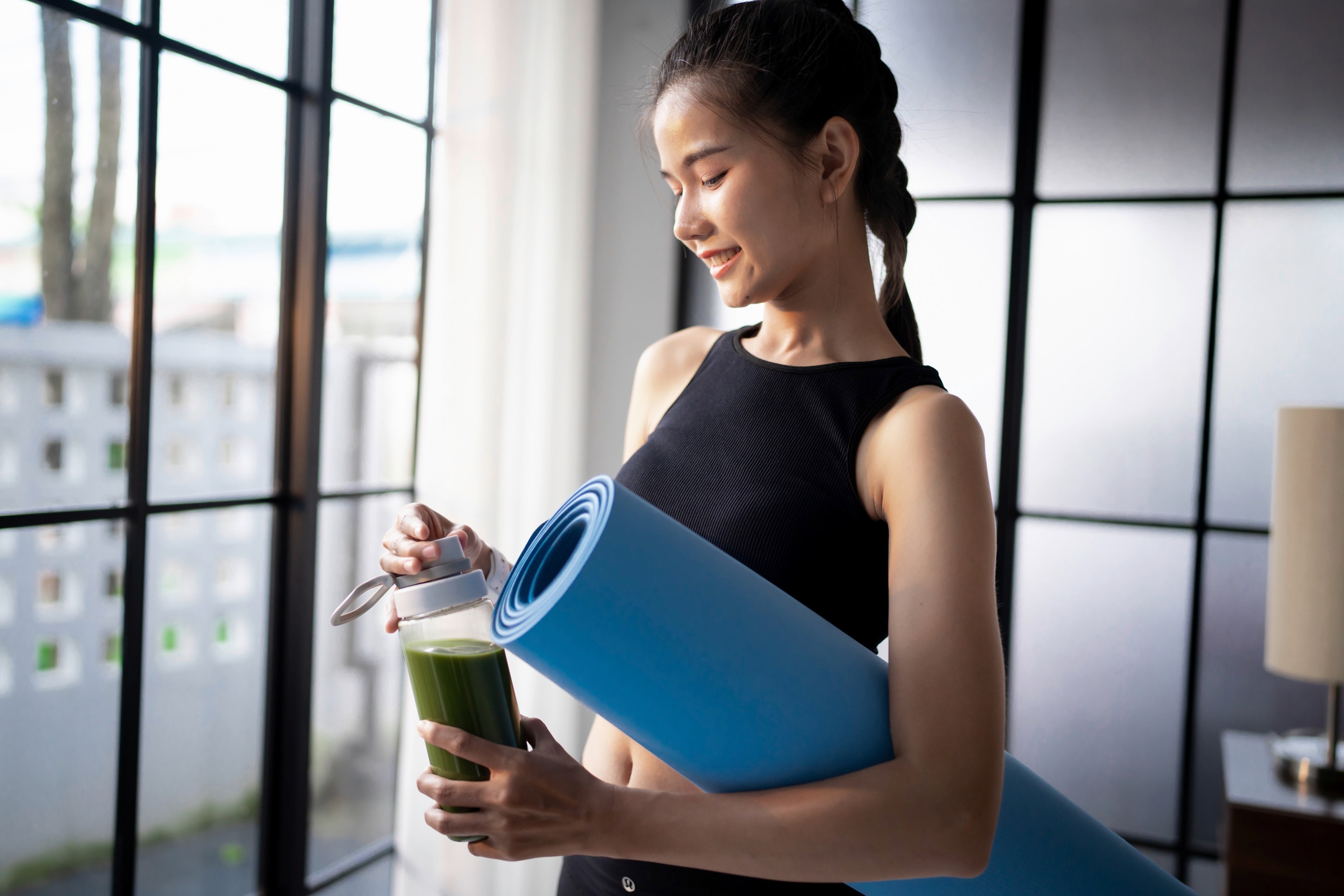 Woman carrying a yoga mat and a green-colored drink.