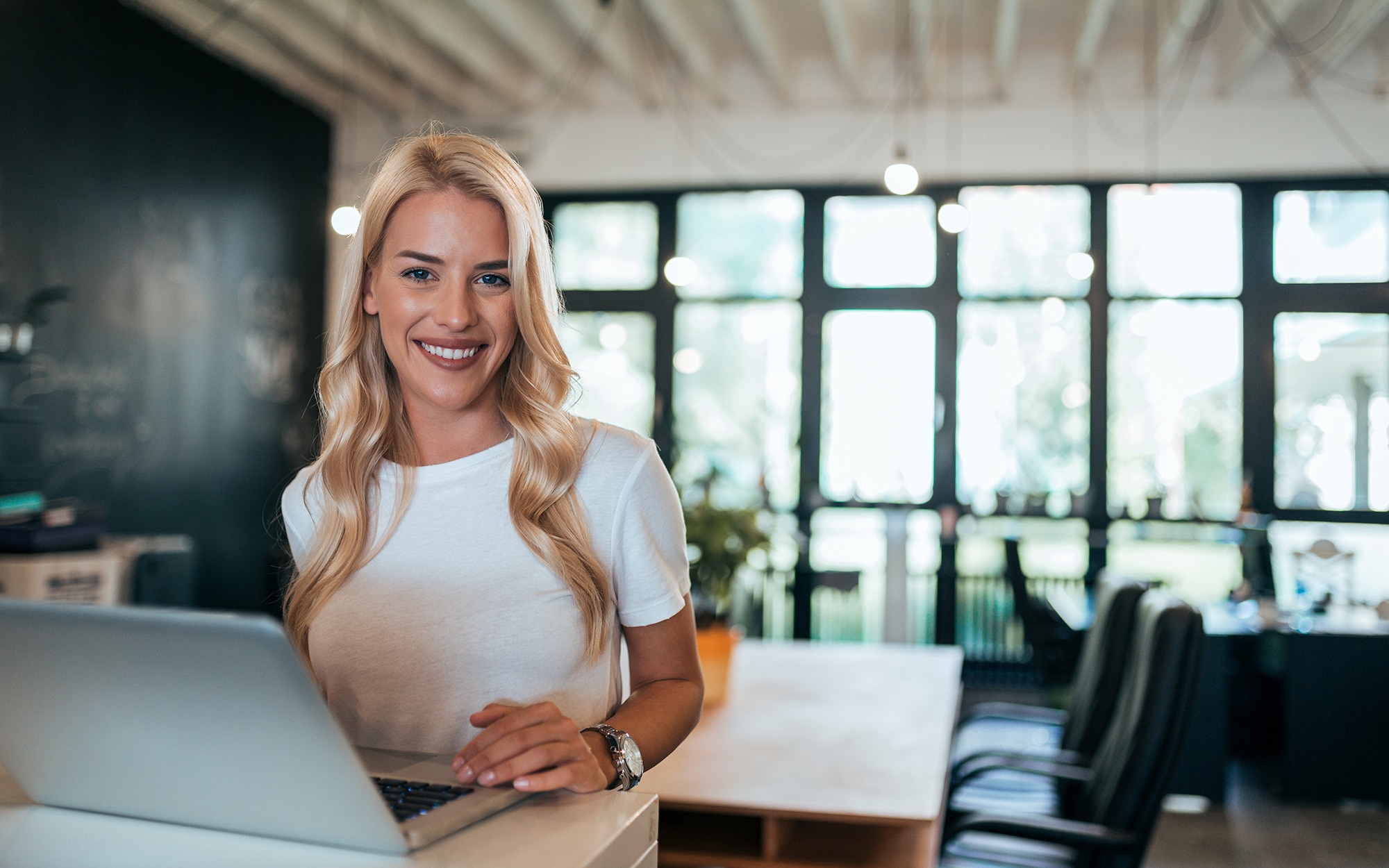 Woman with an easy work hairstyle in an open-plan office setting with window wall in background.