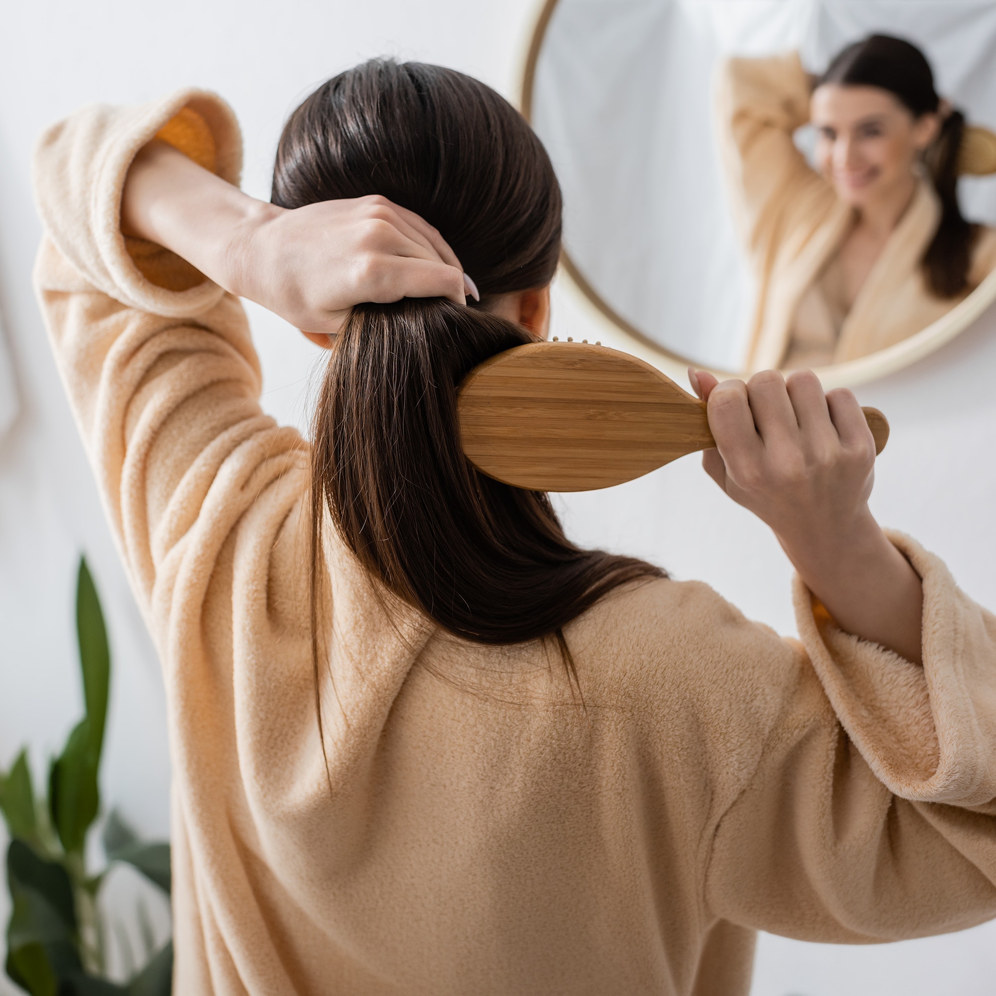 Woman with strong and healthy-looking brown hair brushes her hair while smiling into mirror