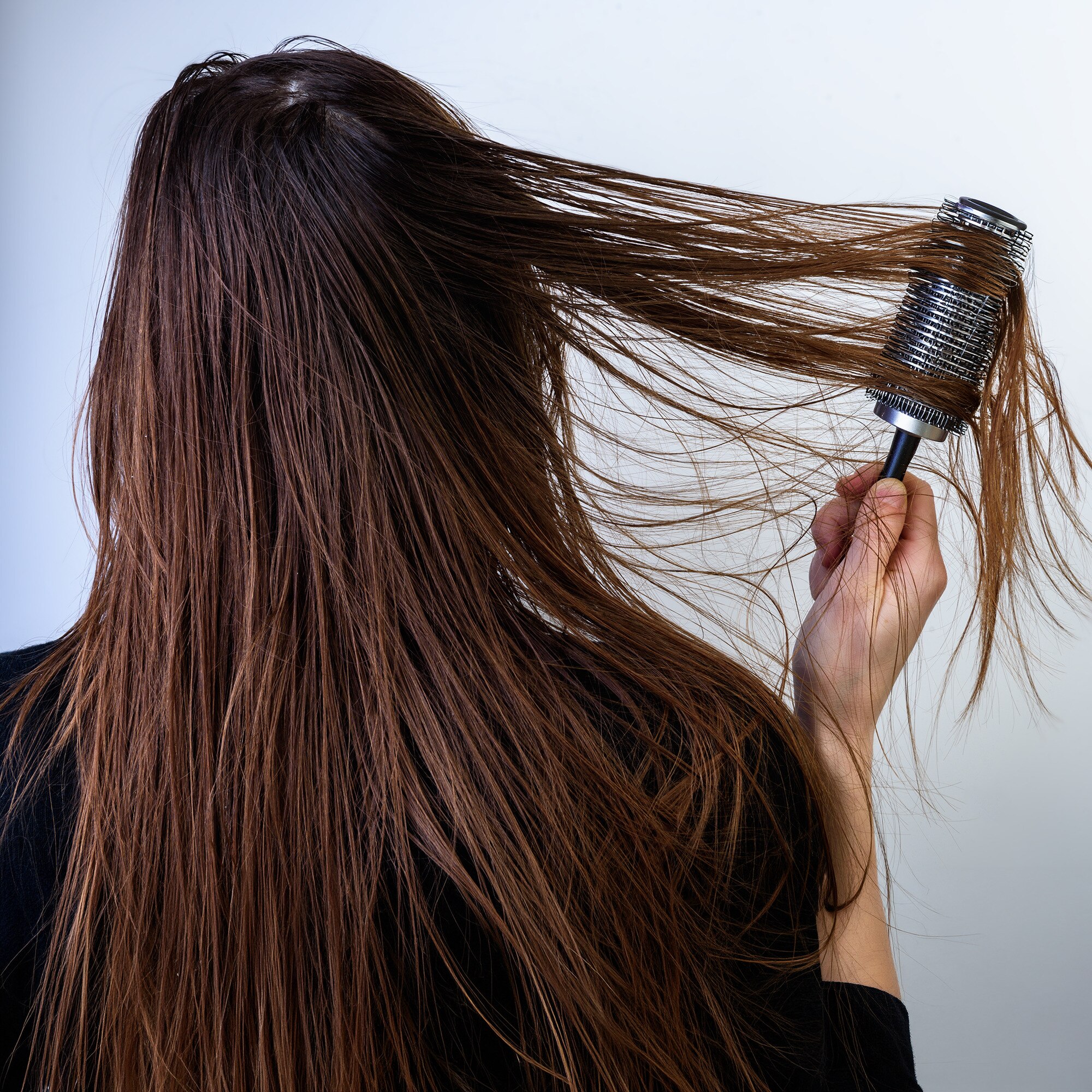 Back view of a woman with long brown hair brushing through her damp hair with a metal roller brush ready to blow dry.