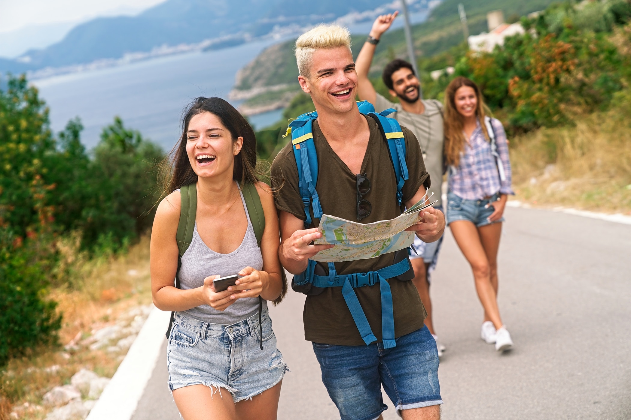 Four young adults laugh while hiking along a scenic coastal road. In the foreground, a woman holds a phone and a man with bleached hair holds a map, while another couple follows behind them.