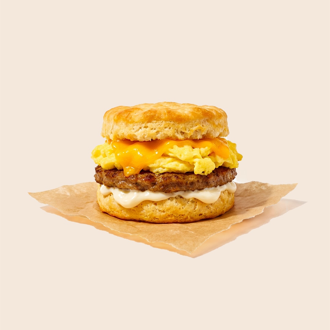 Breakfast sandwich on a biscuit with sausage patty, scrambled eggs, melted cheese, and mayonnaise.