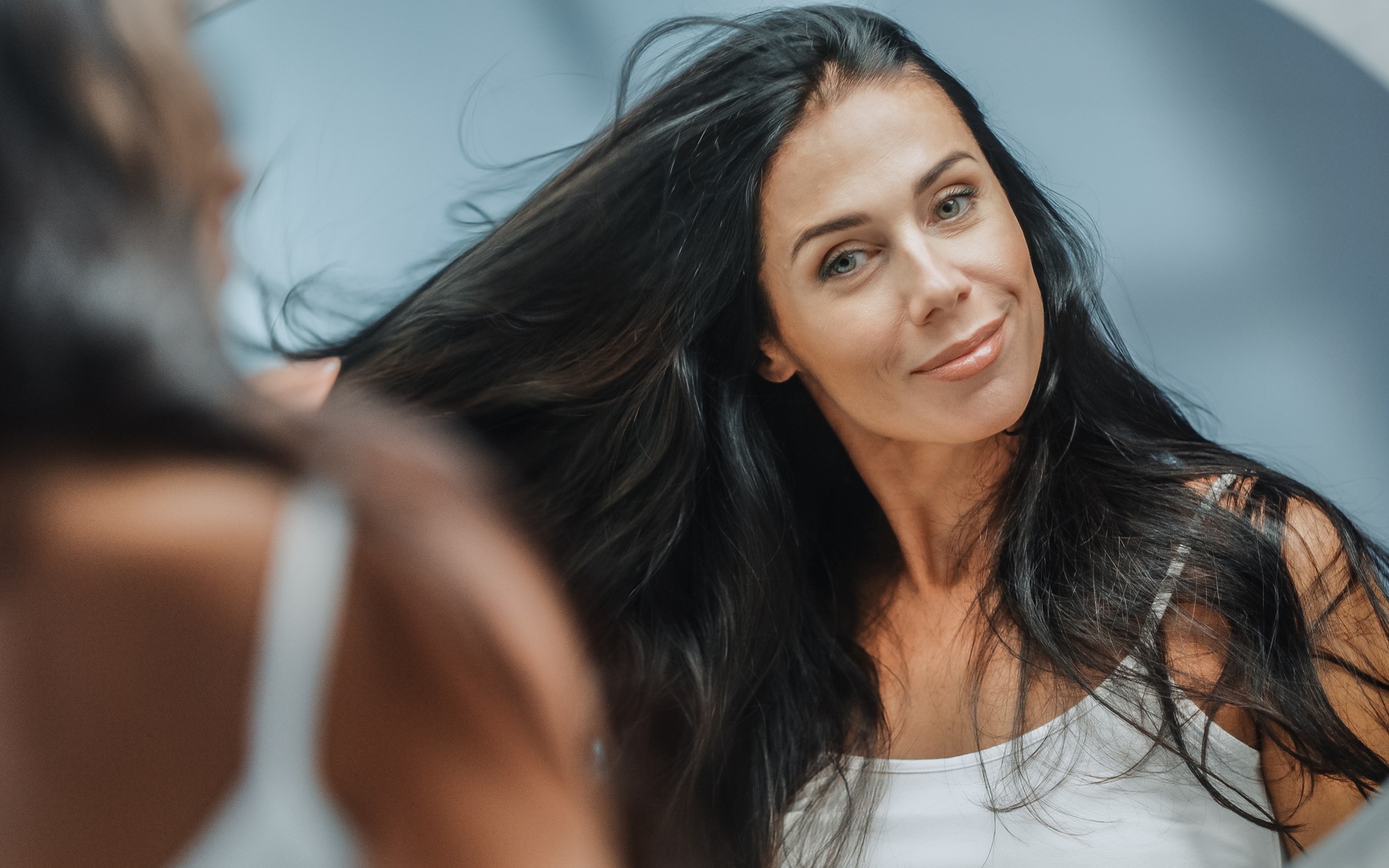 A woman with long, black, layered hair runs a hand through it while smiling in the mirror