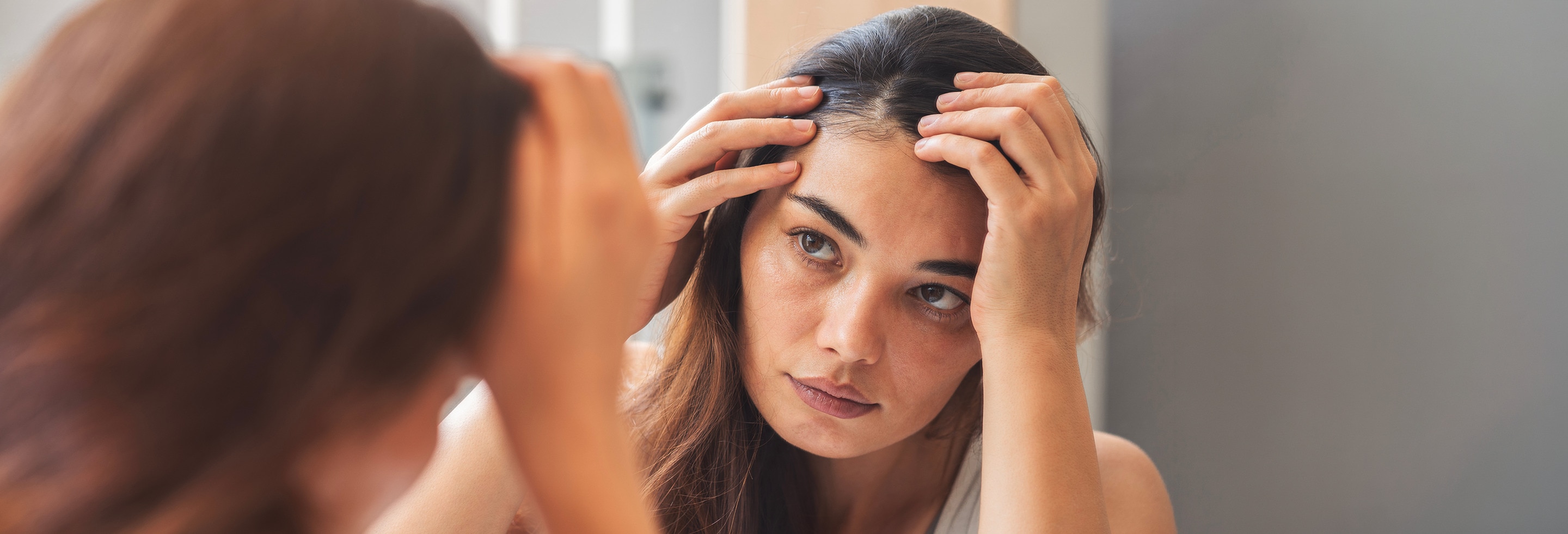 A woman with long, dark hair examines her part line in the mirror.