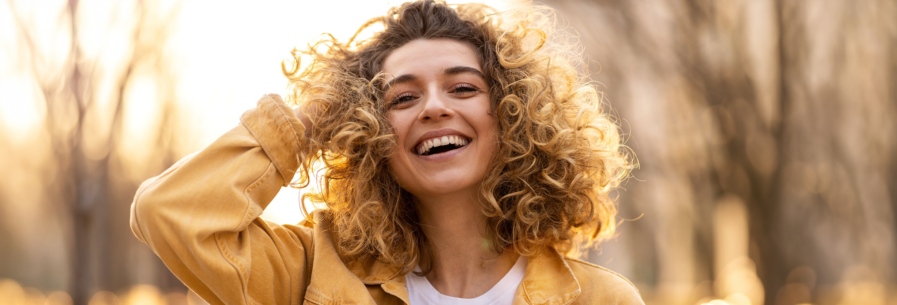 Smiling woman outdoors in late afternoon sun wearing a yellow jacket, touching her curly hair.