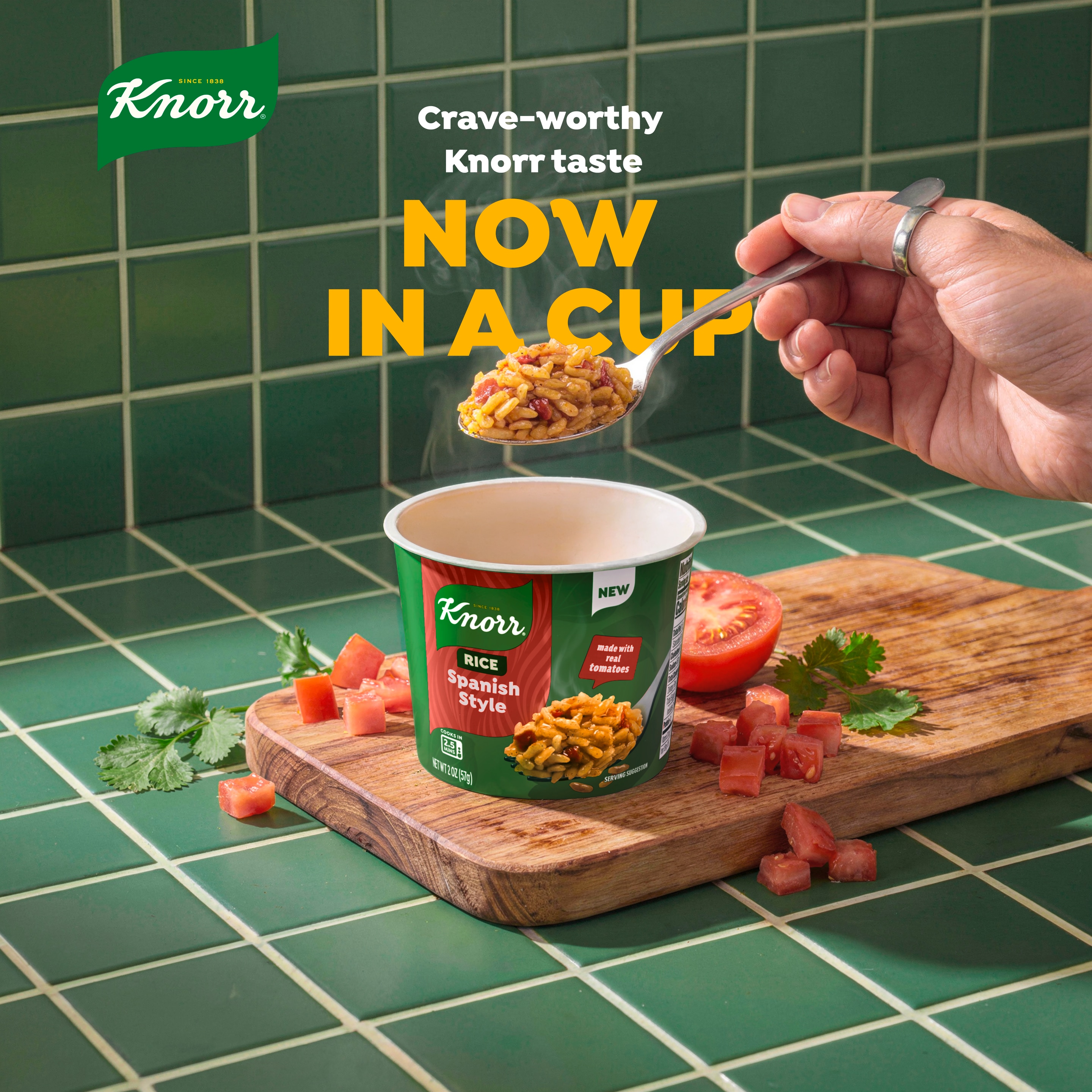 Knorr Spanish Style Rice cup on a wooden cutting board with diced tomatoes and cilantro, while a hand holds a spoonful of steaming rice above the cup.