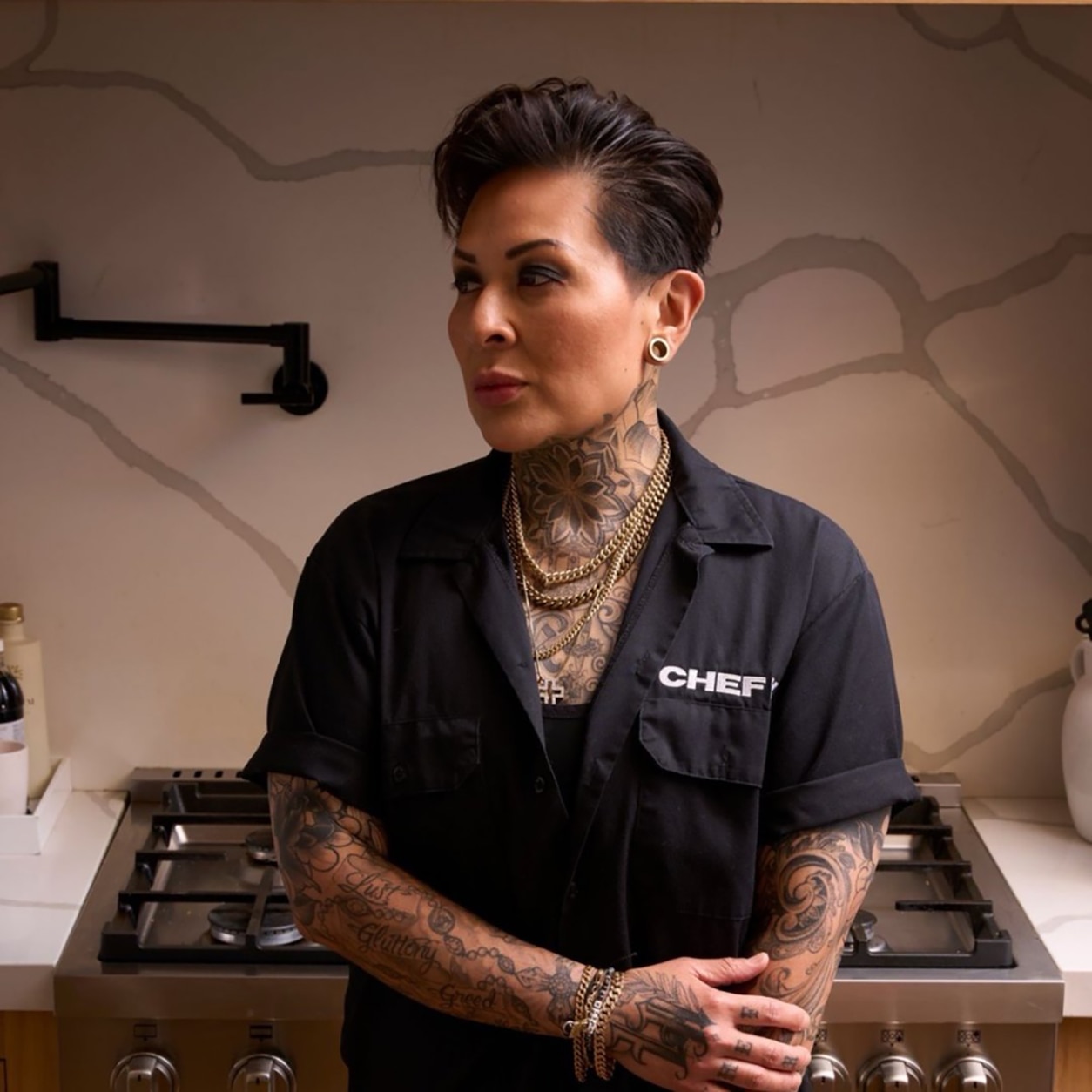 Portrait of a tattooed individual wearing black workwear, standing in a modern kitchen setting with layered jewelry and a contemporary aesthetic.