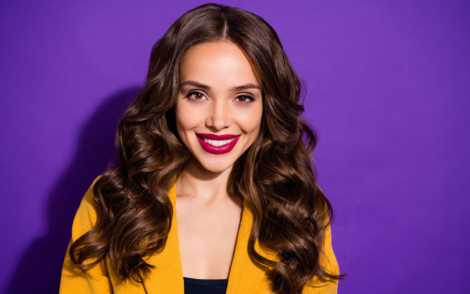 Smartly dressed woman with red lipstick and long, wavy, glossy brown hair against a vivid purple background