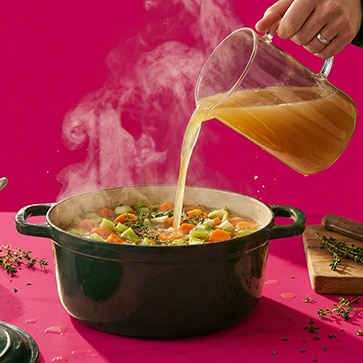 Broth being poured into a pot of vegetable soup on a bright pink background