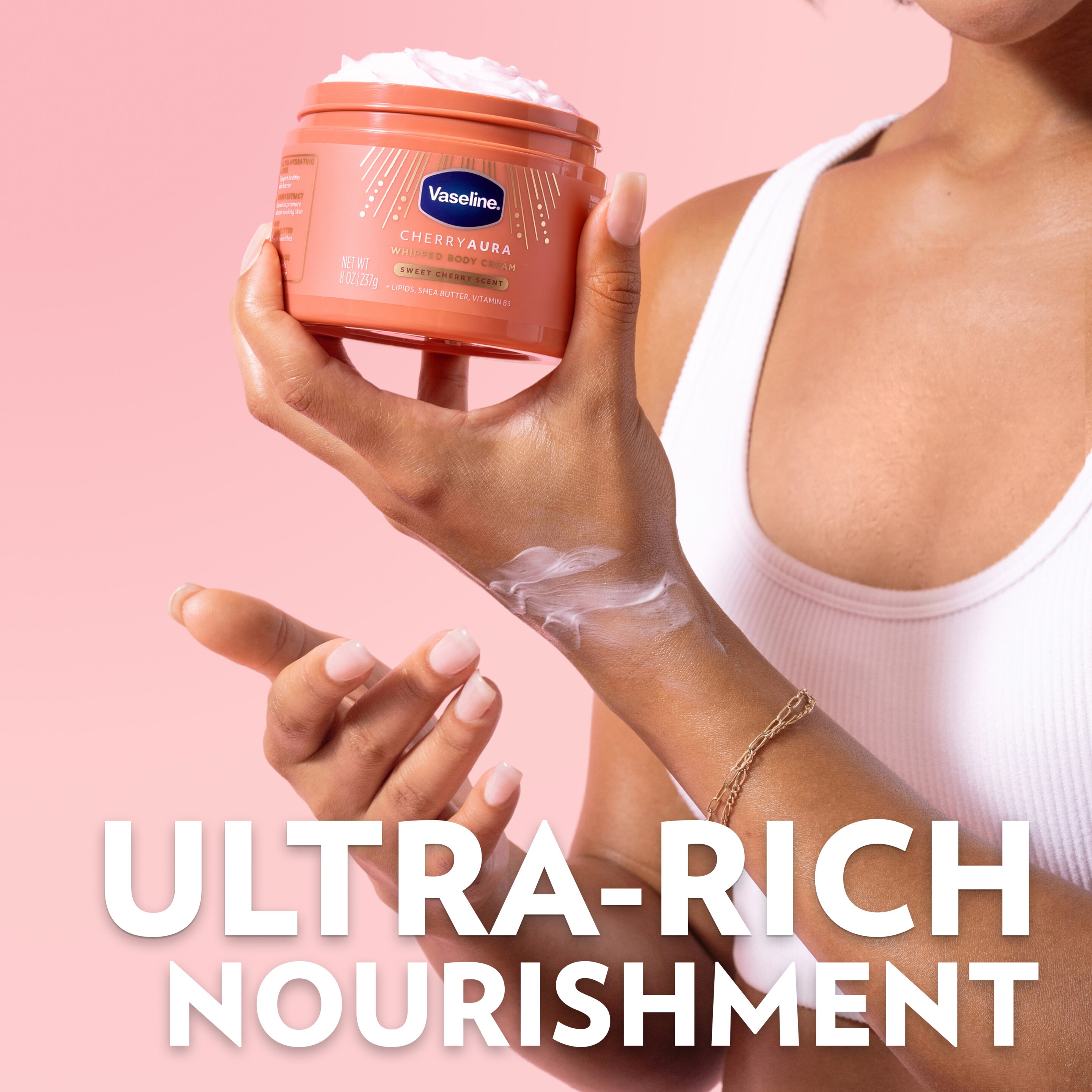 A person holds a jar of Vaseline Cherry Aura Whipped Body Cream with a swipe of cream on the hand, alongside text