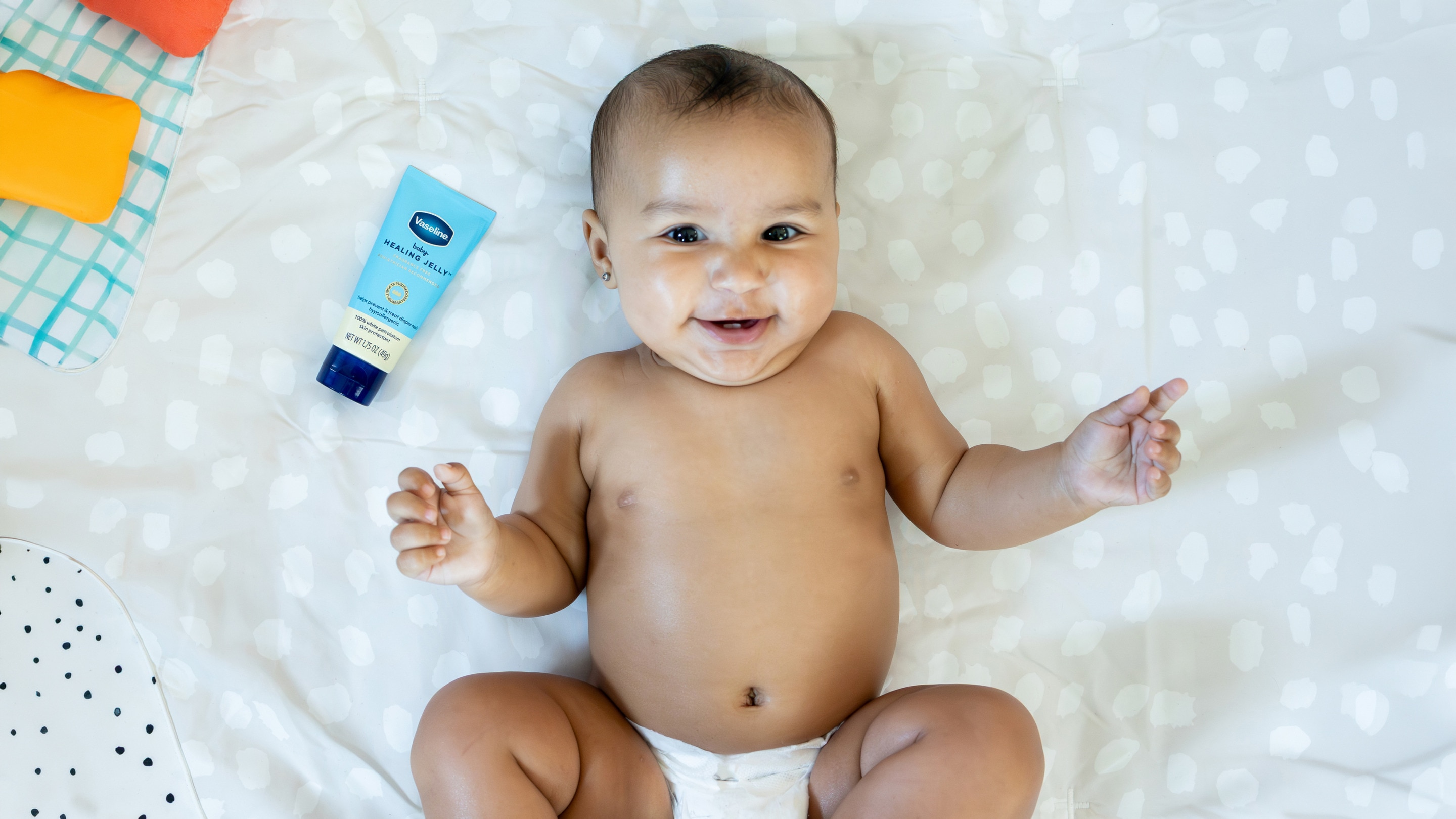 Baby wearing a diaper lying on a white patterned blanket next to a tube of Vaseline Healing Jelly.