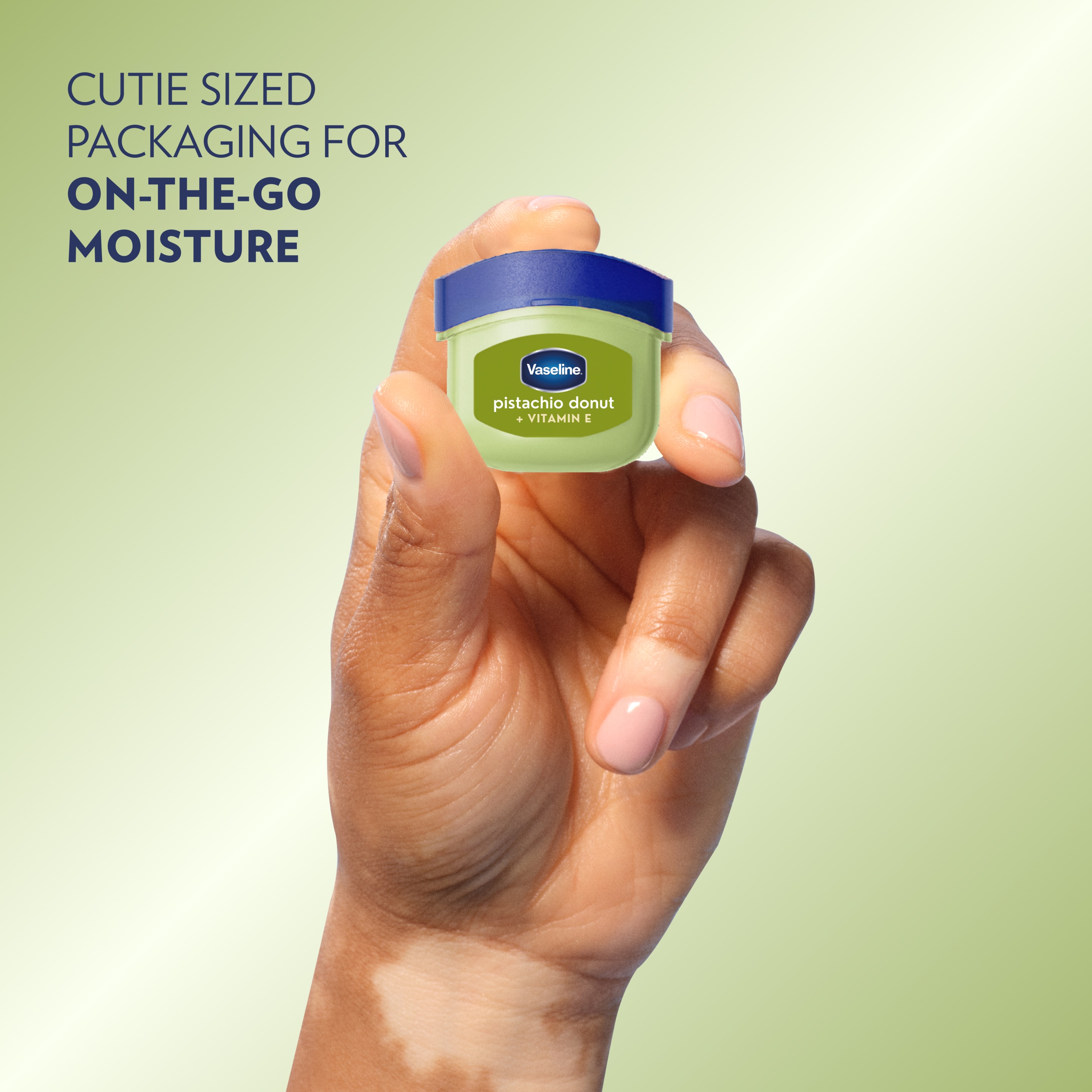 Close-up of a hand holding a small container of Vaseline Pistachio Donut + Vitamin E lip balm against a light green background, with text
