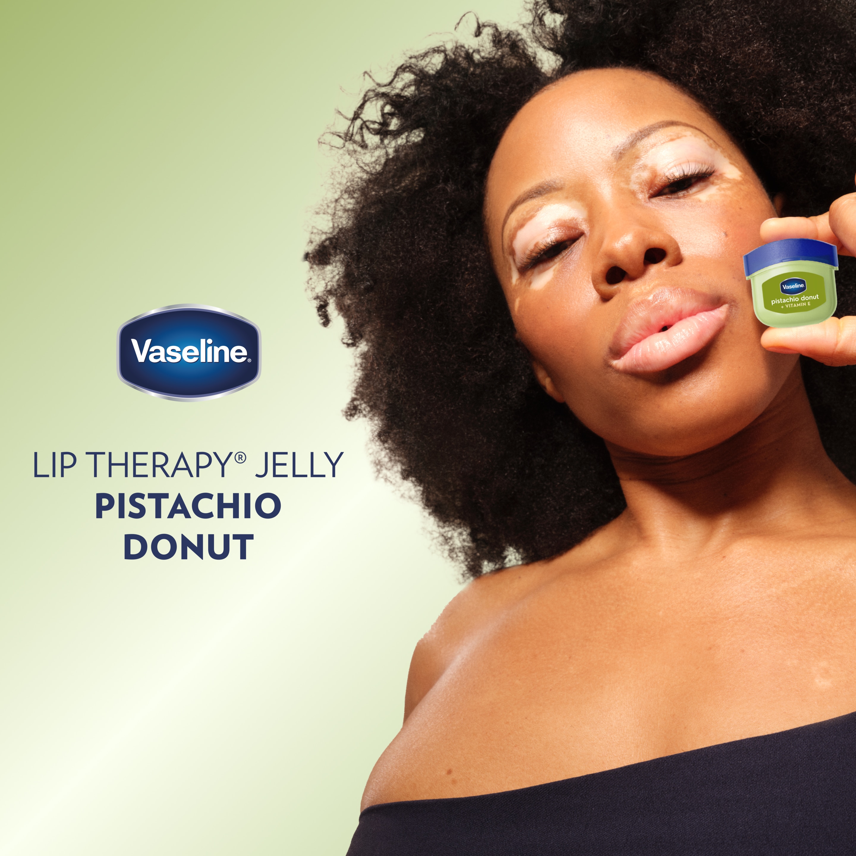 A person holding a small container of Vaseline Lip Therapy Jelly Pistachio Donut near their face against a light green background, with the Vaseline logo and text