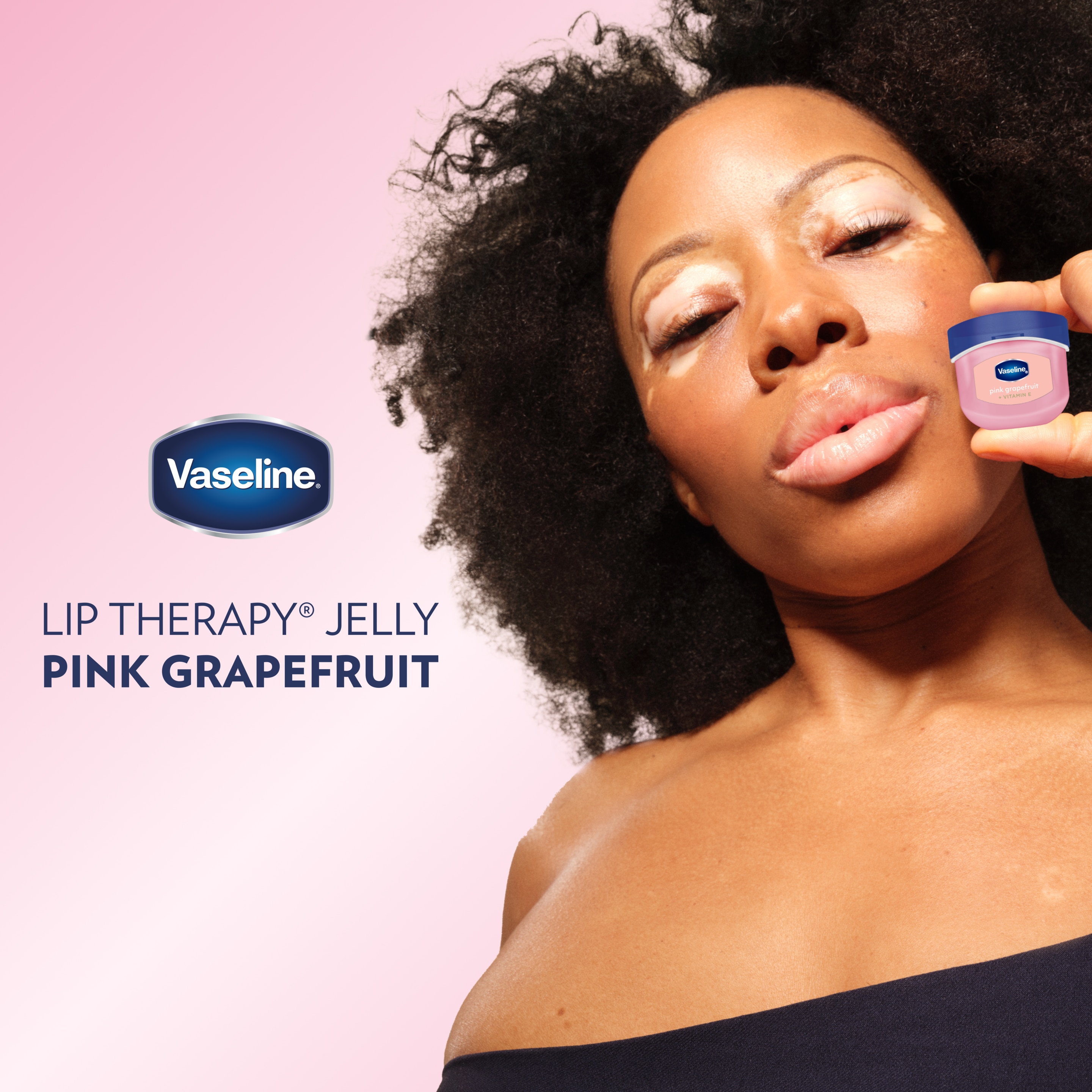 A person holding a small container of Vaseline Lip Therapy Jelly Pink Grapefruit near their face against a pink background, with the Vaseline logo text