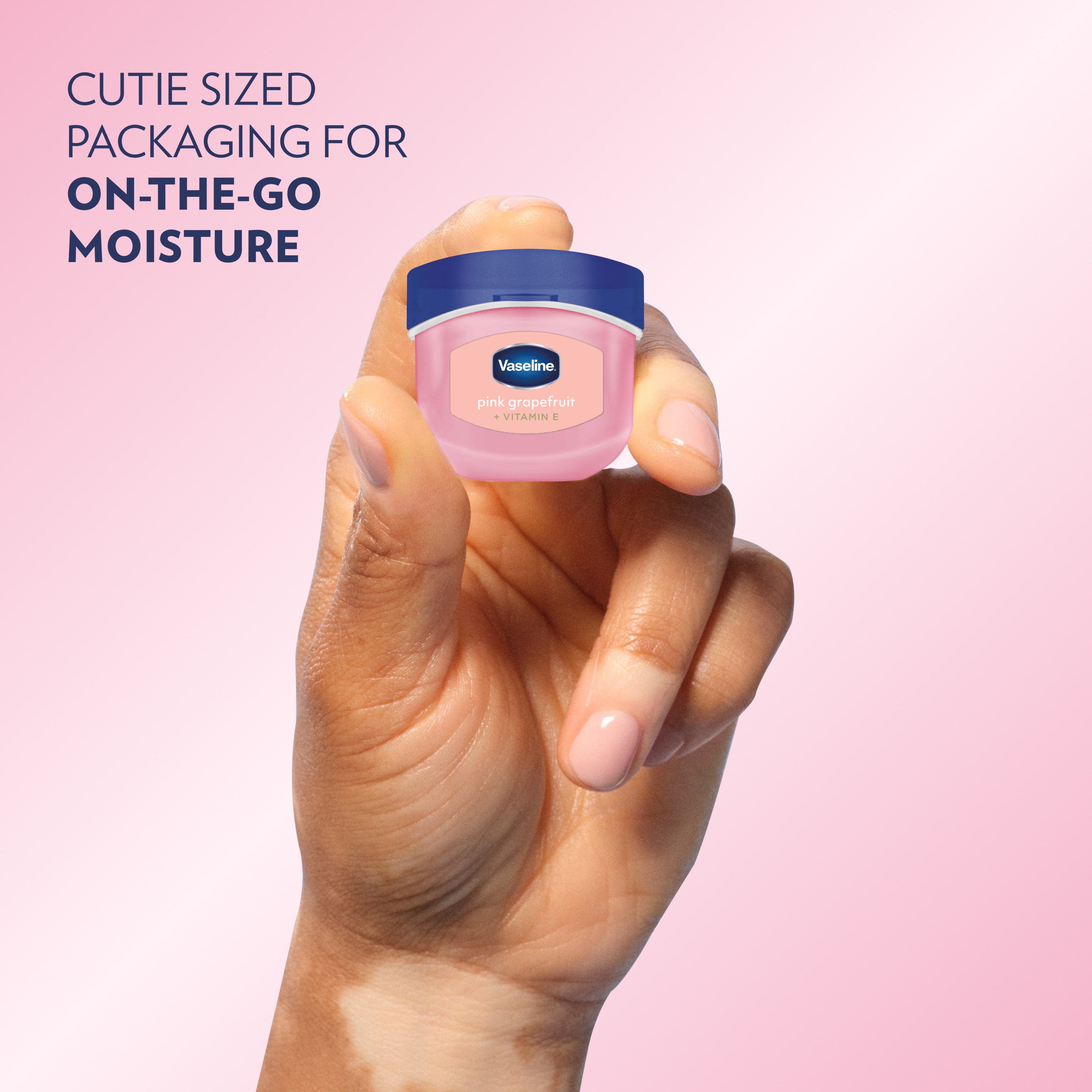 Close-up of a hand holding a small container of Vaseline Pink Grapefruit + Vitamin E lip balm against a pink background, with text